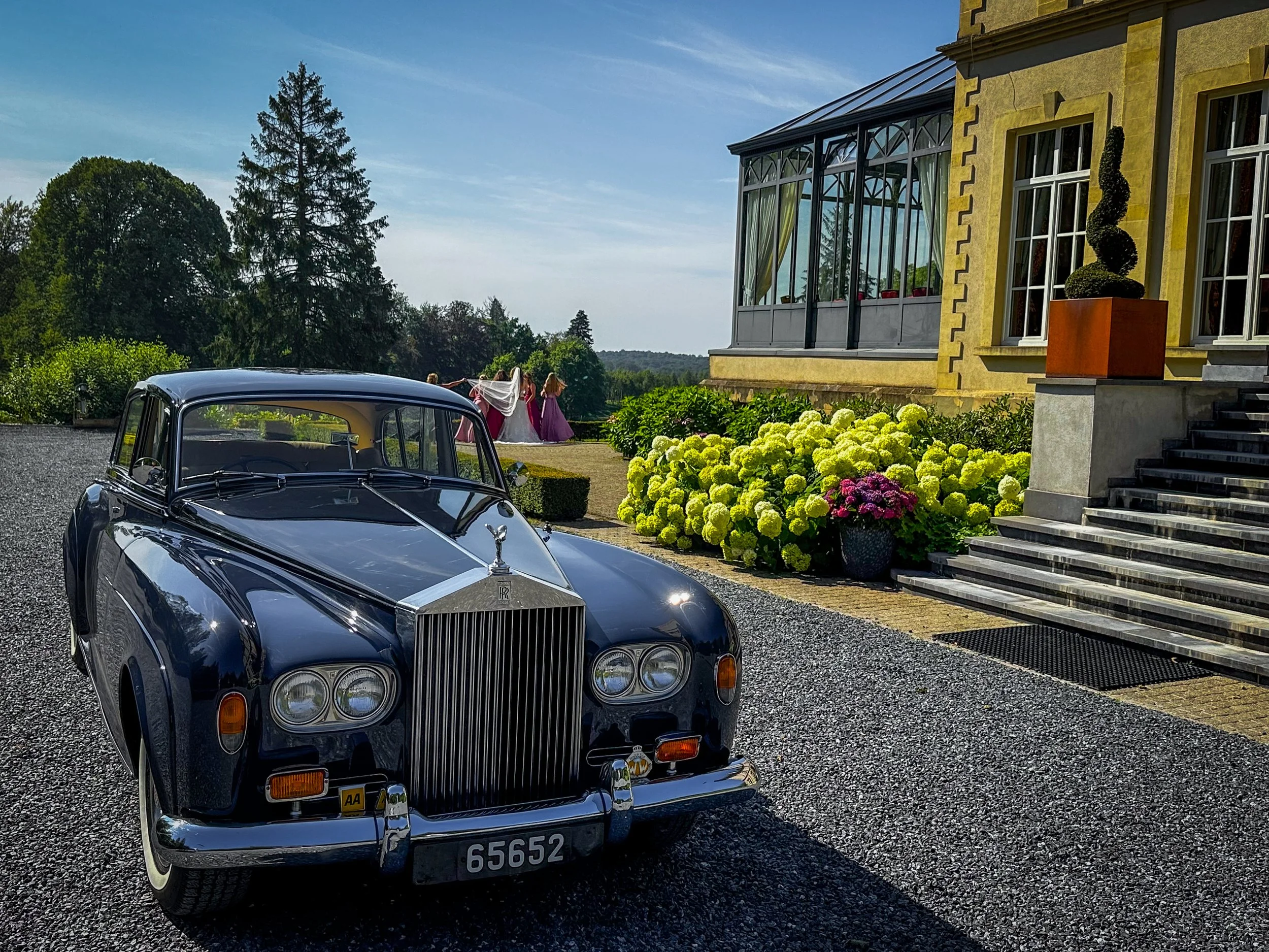 Une voiture classique noire est garée devant une maison élégante avec un escalier en pierre, des buissons fleuris et des sculptures de topiaires, dans un cadre verdoyant en plein air, lors d'une journée ensoleillée.