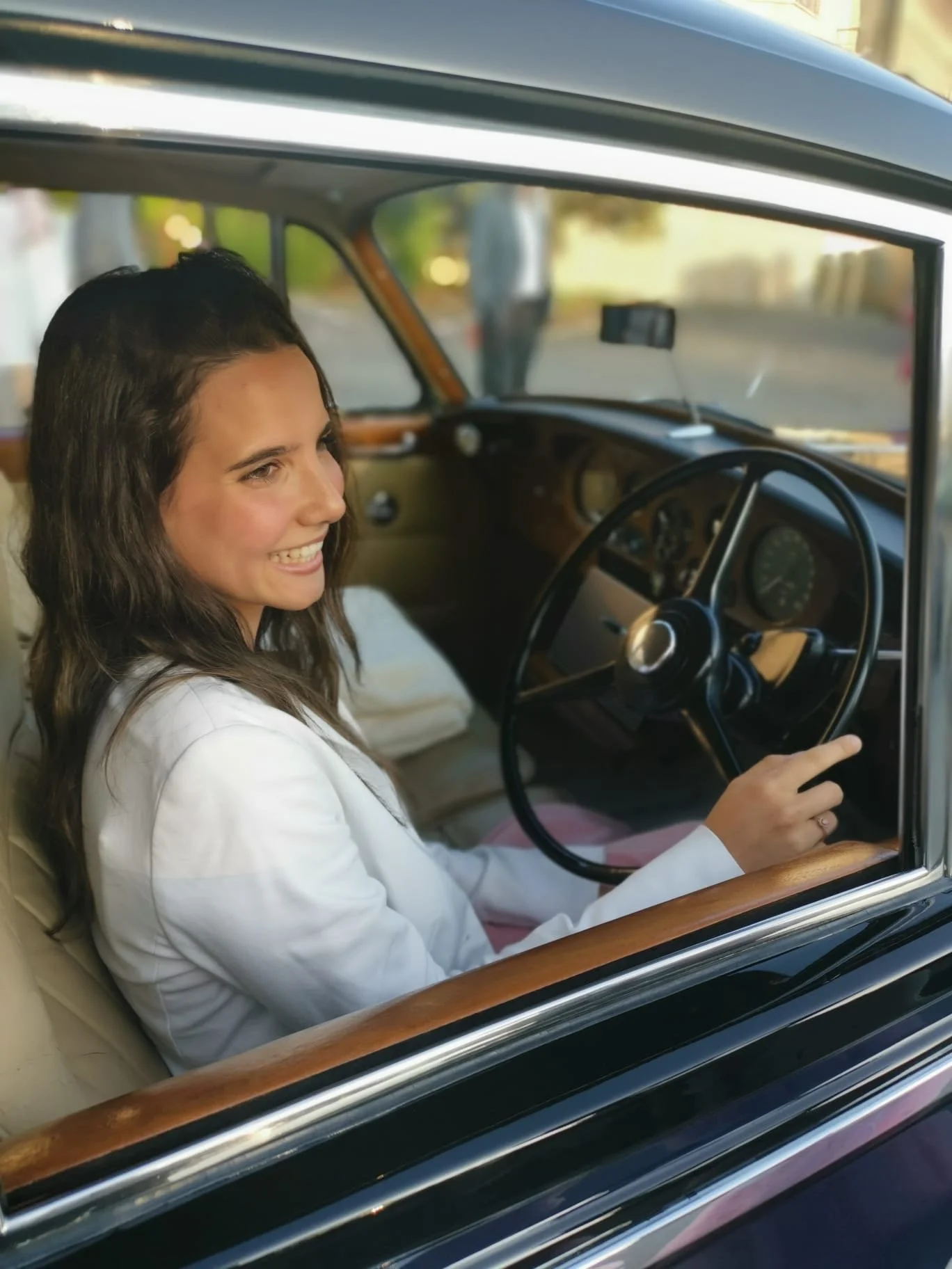 Une femme souriante assise dans une voiture vintage, vue de l'extérieur à travers la fenêtre