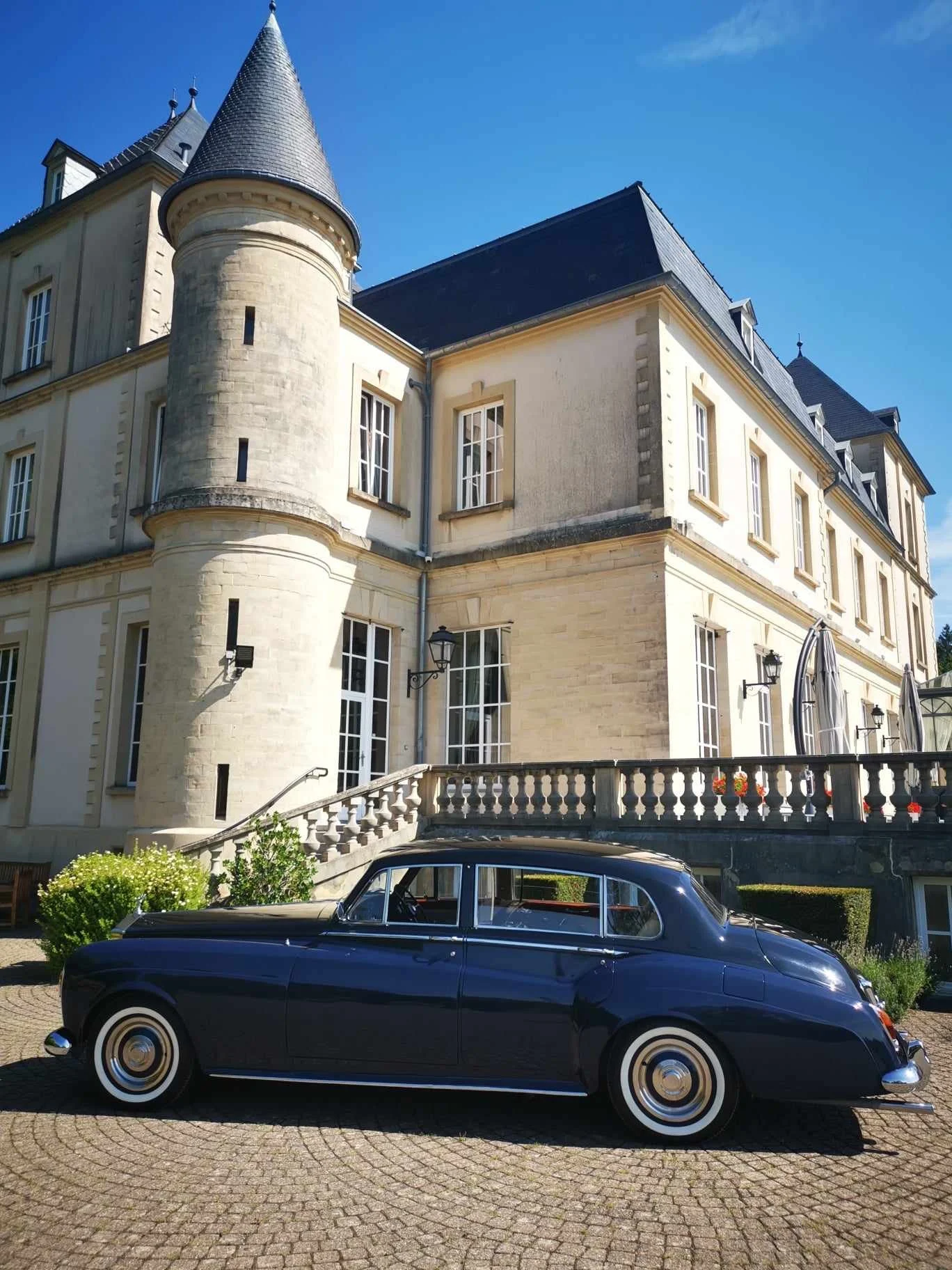 Une voiture ancienne noire stationnée devant un château de style médiéval avec une tour ronde, sous un ciel bleu clair.
