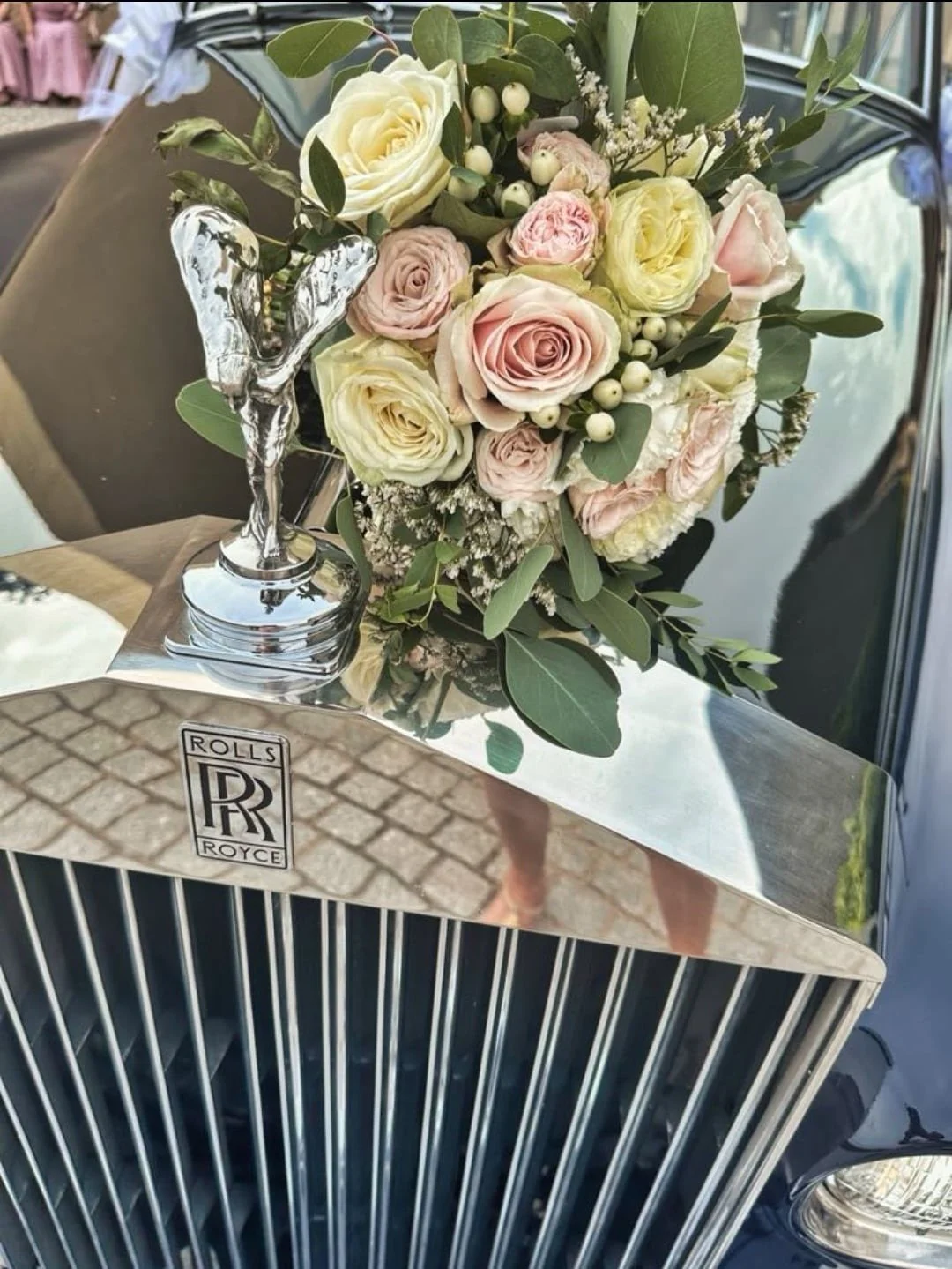 Un bouquet de fleurs roses, blanches et crème avec des feuilles vertes, placé sur la calandre d'une voiture Rolls Royce, avec un petit trophée argenté en forme de papillon à côté.
