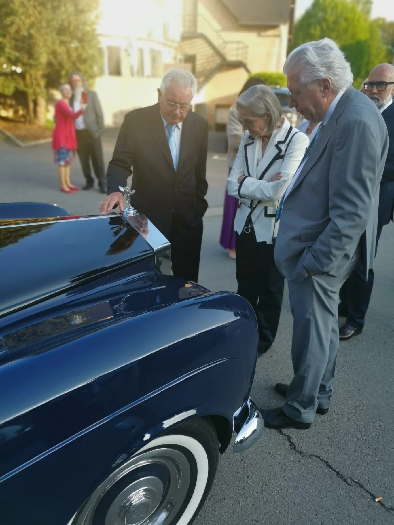 Groupe de personnes en vêtements formels regardant une voiture ancienne noire, probablement lors d'un événement ou d'une rencontre sociale, dans une rue résidentielle en plein air.