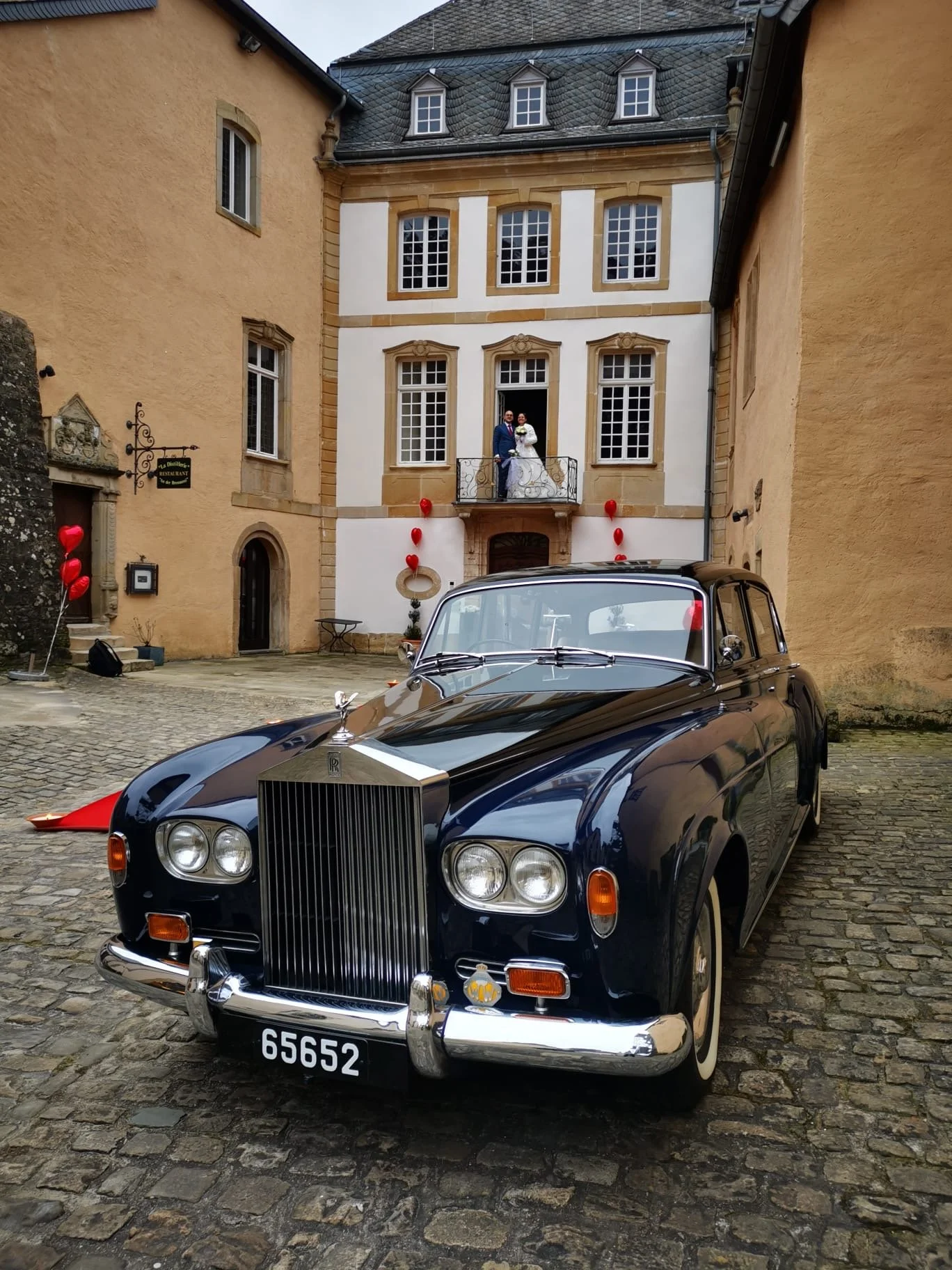 Une voiture ancienne noire garée dans une cour pavée, avec un couple de mariés sur un balcon d'un bâtiment historique en arrière-plan, décoré de ballons en forme de cœur.