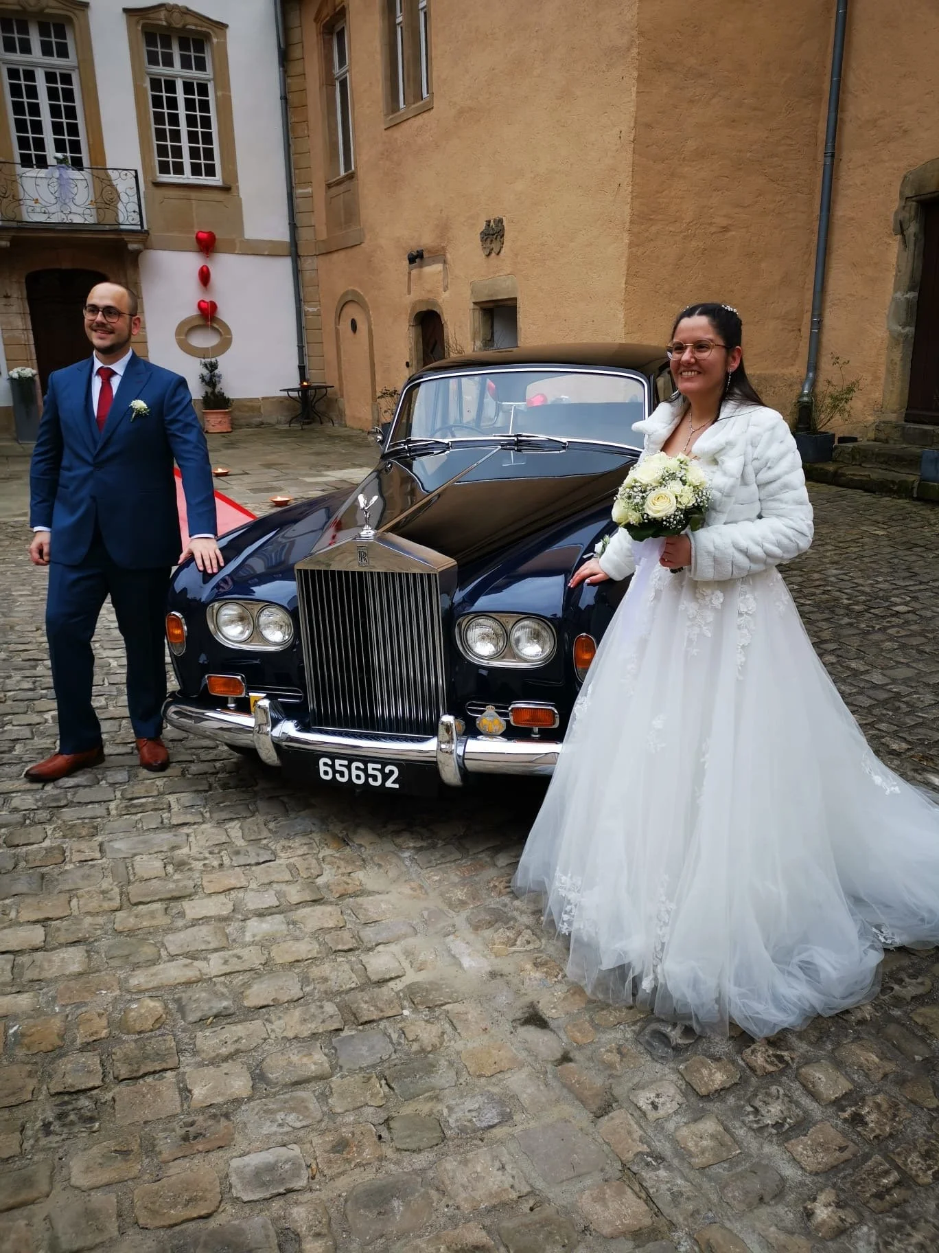 Un couple marié, un homme en costume bleu et une femme en robe de mariée blanche, posant à côté d'une voiture ancienne noire dans une cour pavée, décorée avec des ballons en forme de cœur rouges, pour leur mariage.