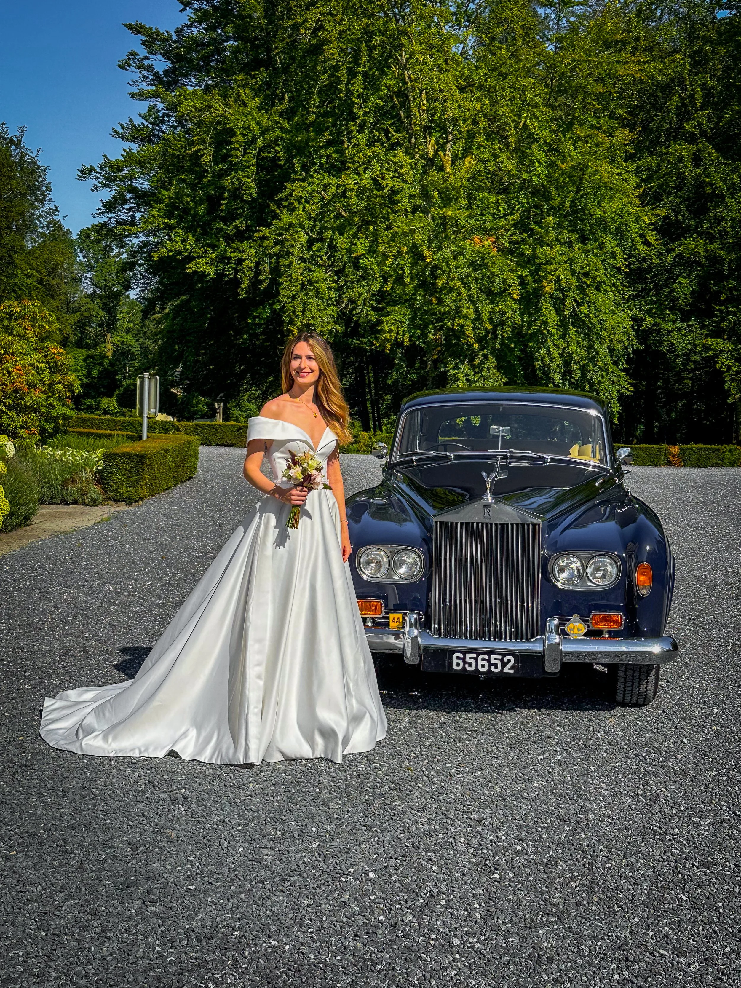 Une jeune femme en robe de mariage blanche, tenant un bouquet de fleurs, se tenant à côté d'une voiture ancienne noire sur une route entourée d'arbres verts.