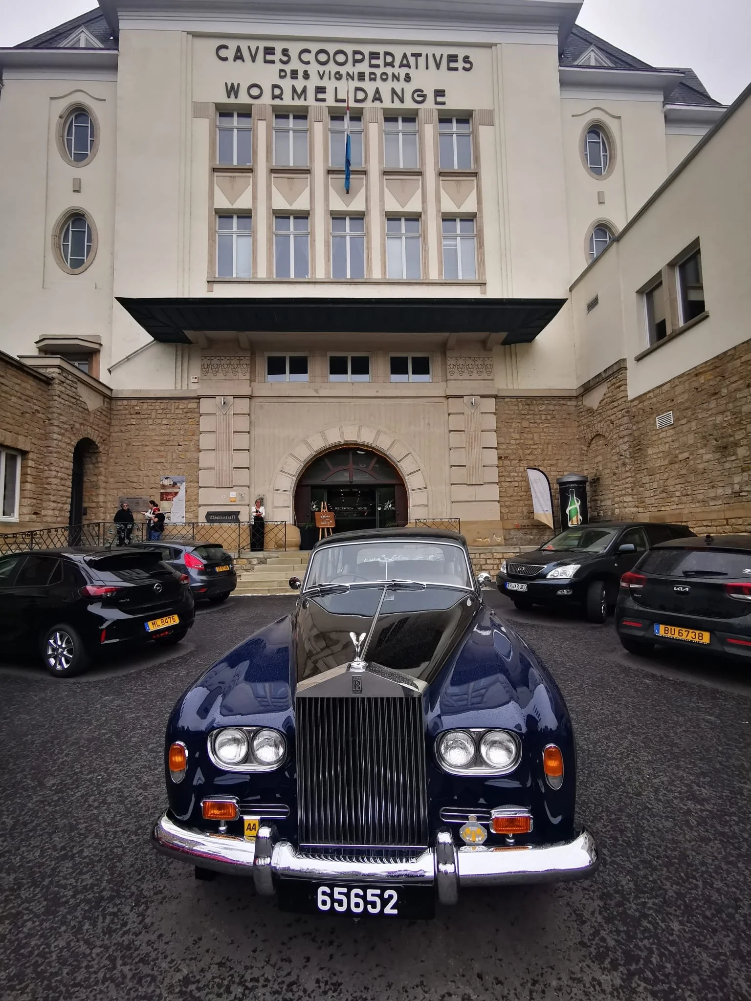 Une voiture classique Rolls-Royce noire garée devant un bâtiment avec un panneau indiquant CAVES COOPERATIVES DES VIGNERONS WORMELDANGE.