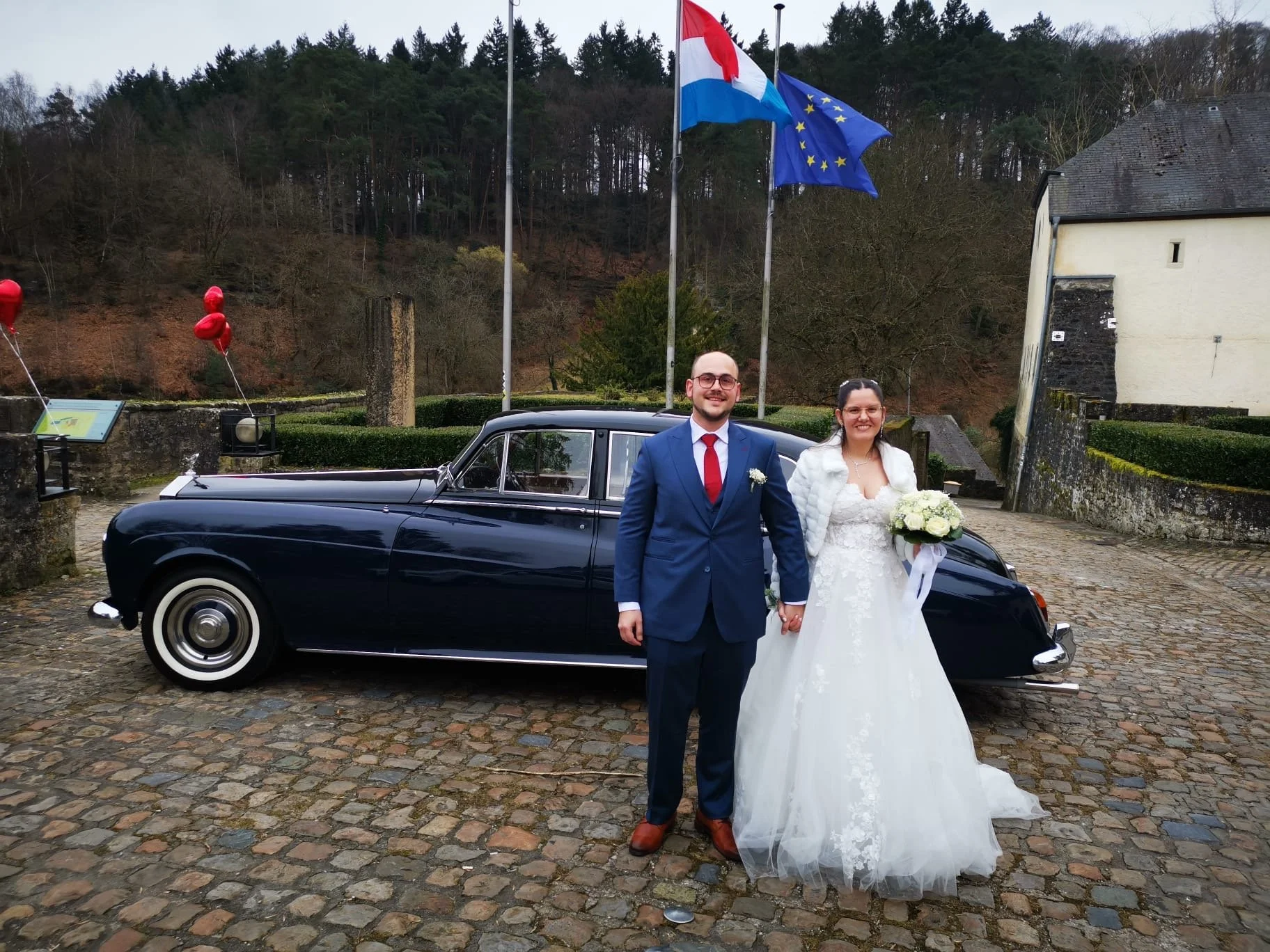 Un couple de mariés posant devant une voiture ancienne noire sur une place pavée, avec des drapeaux français et européens en arrière-plan.