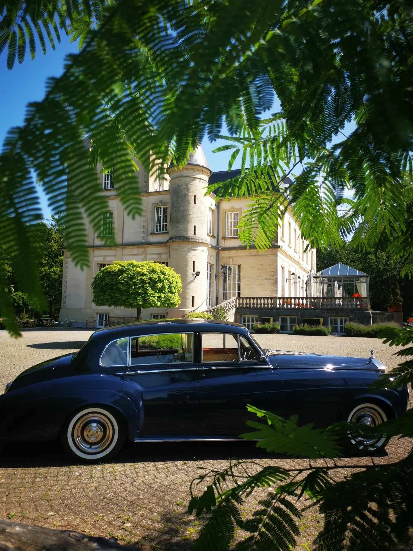Une voiture classique noire garée devant un château avec une terrasse et des fenêtres anciennes, entouré de verdure et des arbres.