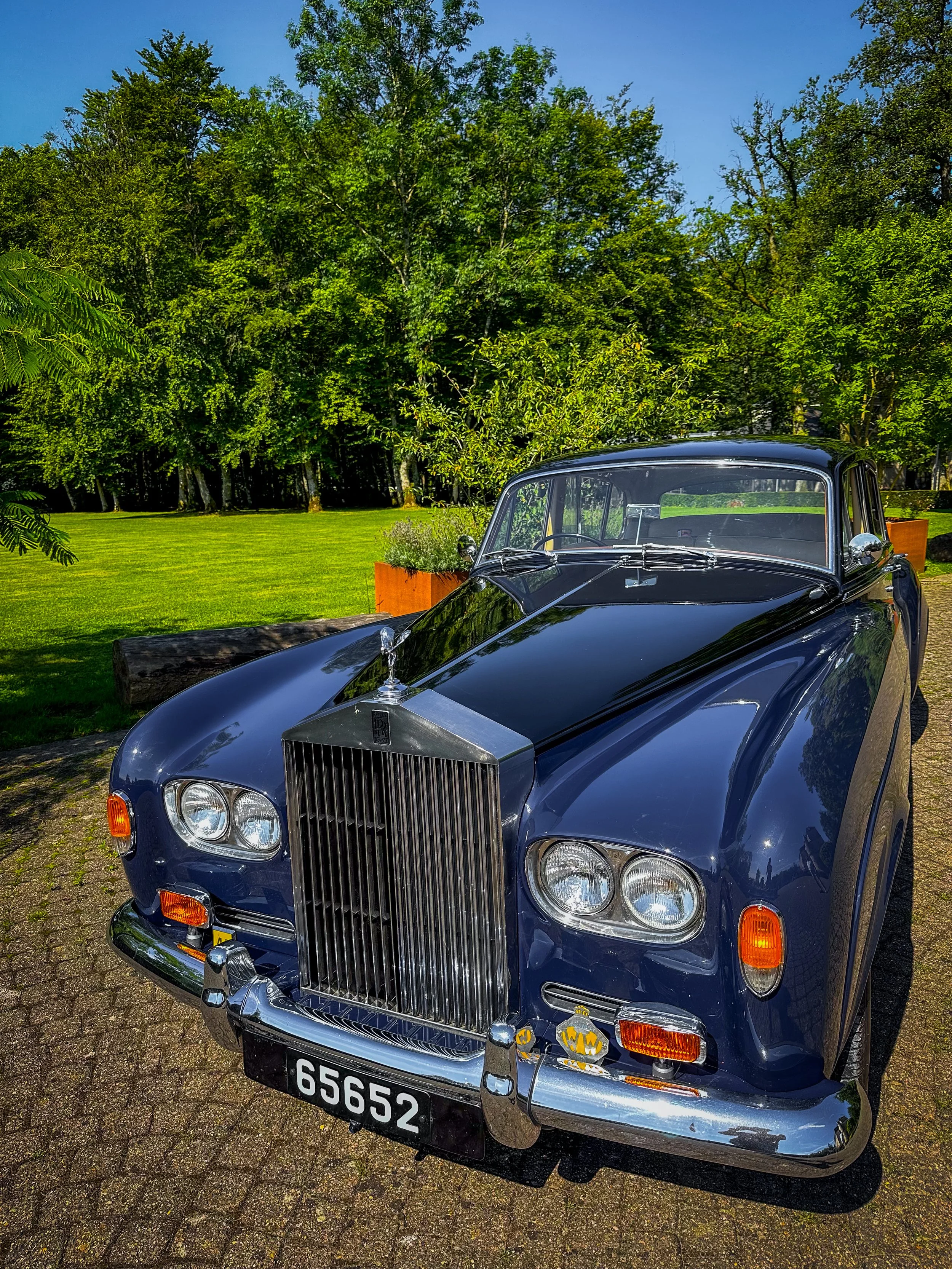 Une voiture de collection noire avec un radiateur vertical chromé, garée sur un chemin pavé, avec un fond de verdure et d'arbres sous un ciel bleu.