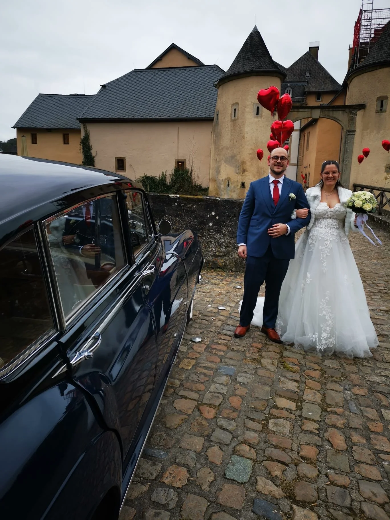 Un couple de mariés posant devant une voiture noire devant un château avec des ballons en forme de cœur rouges, lors d'un mariage.