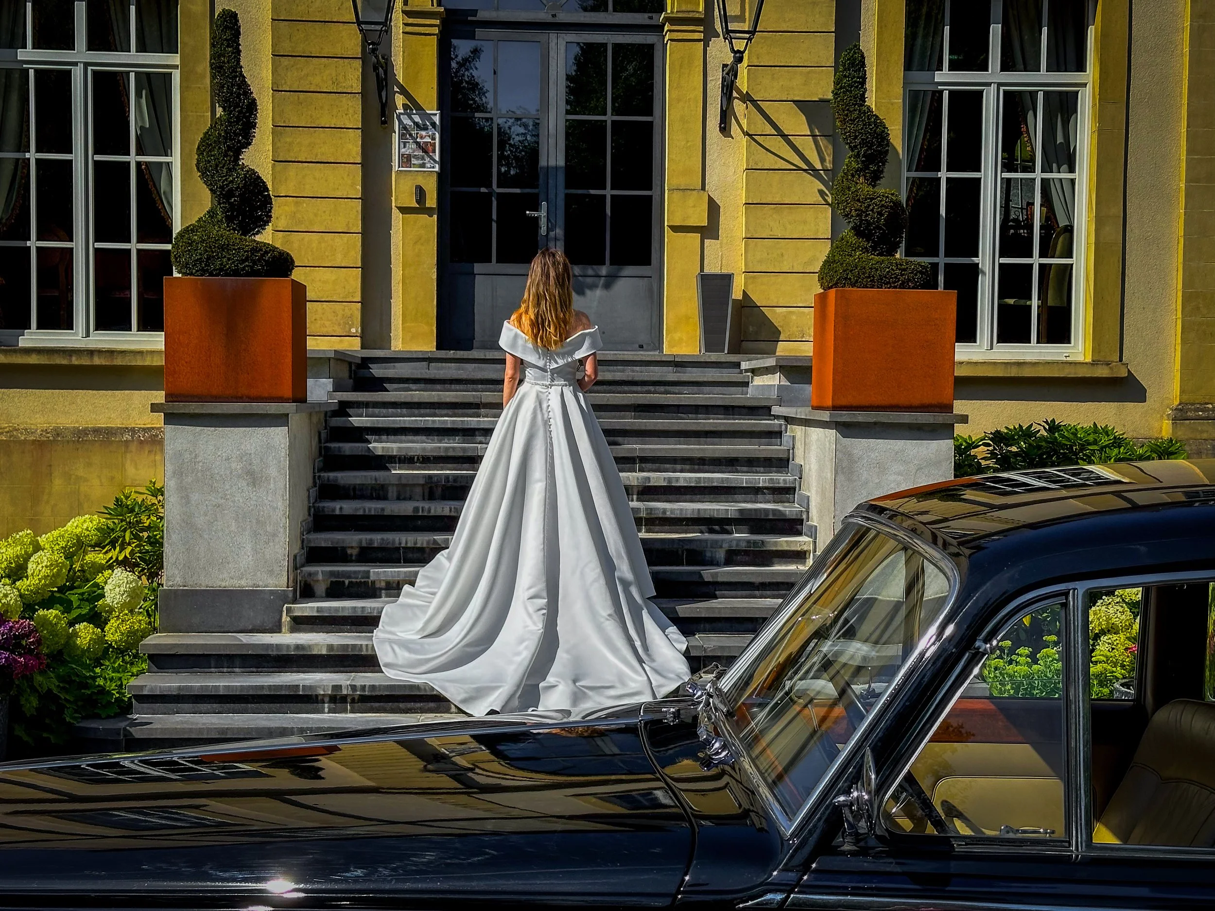 Une femme en robe de mariage blanche se tient devant une maison jaune, vue de dos, avec une voiture noire au premier plan.