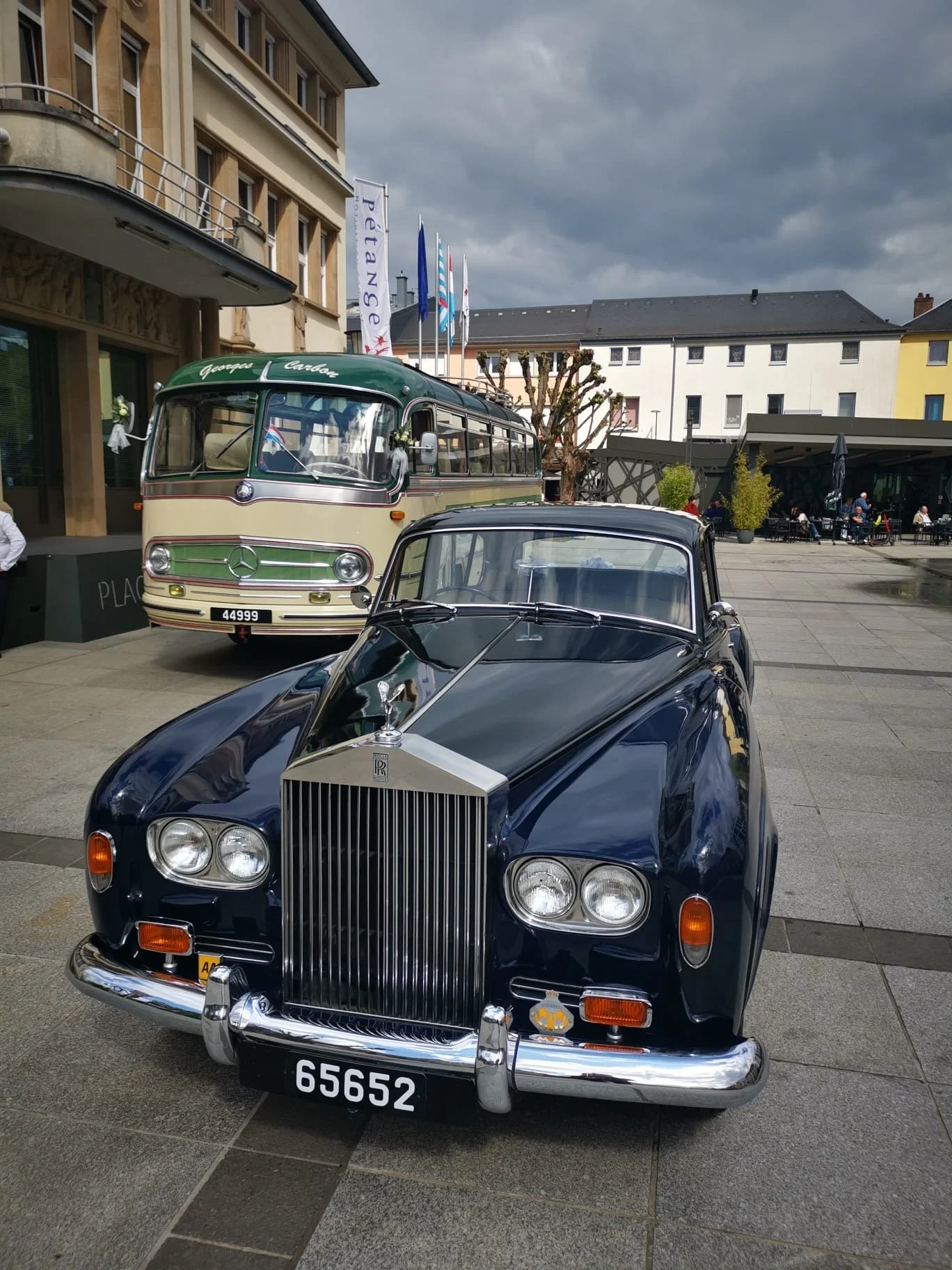Une voiture Rolls-Royce classique noire exposée en plein air, avec un autobus vintage Mercedes-Benz derrière, dans une place urbaine. Ciel nu et bâtiments modernes en arrière-plan.