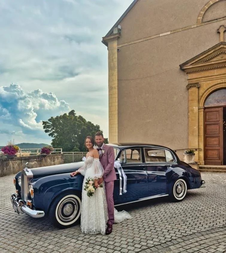 Un couple de mariés posant devant une voiture classique noire à l'extérieur d'une église, avec un ciel nuageux en arrière-plan.
