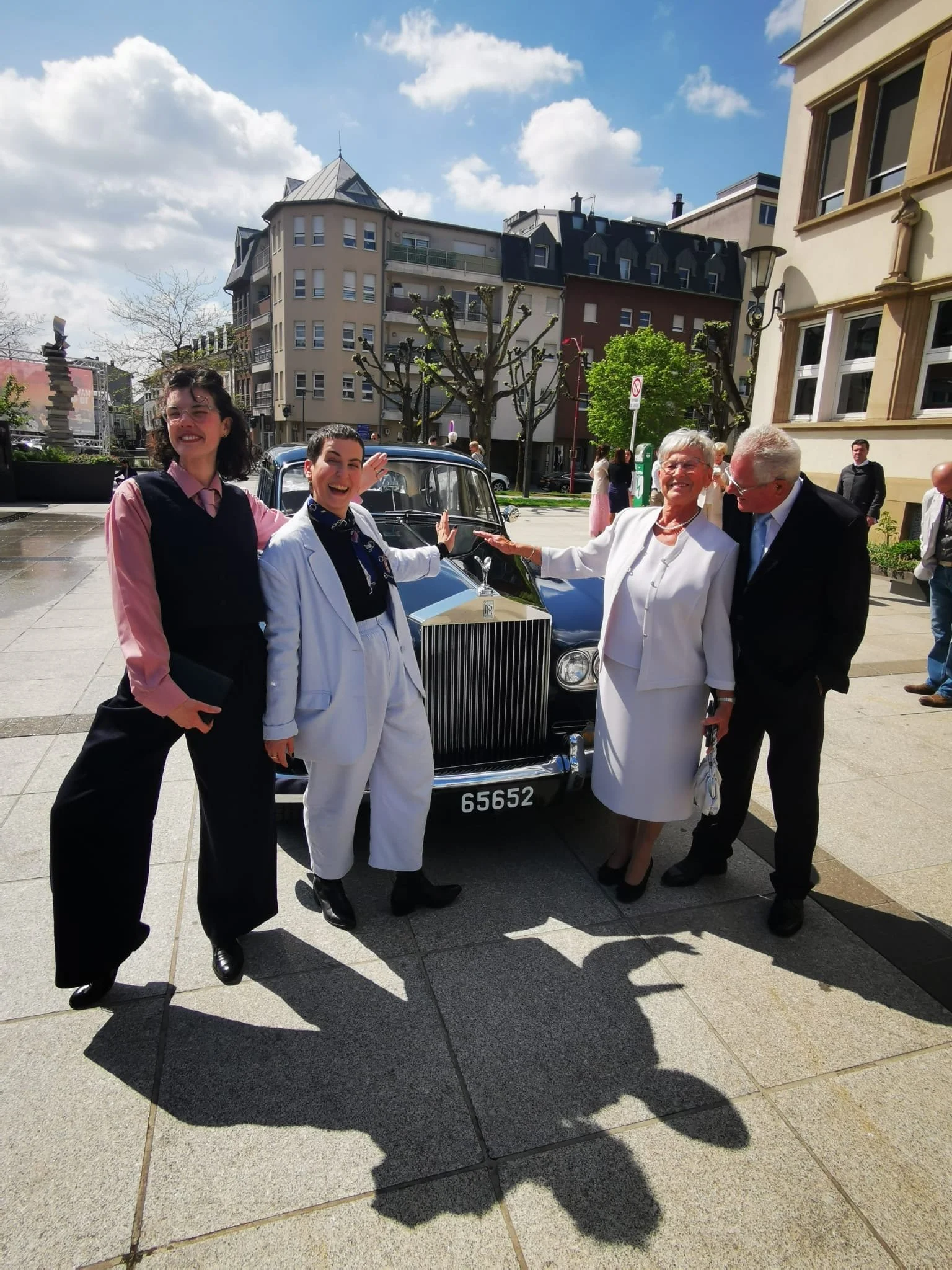 Groupe de cinq personnes souriantes posant devant une voiture classique noire, en plein air sous un ciel en partie nuageux, avec des bâtiments résidentiels en arrière-plan.