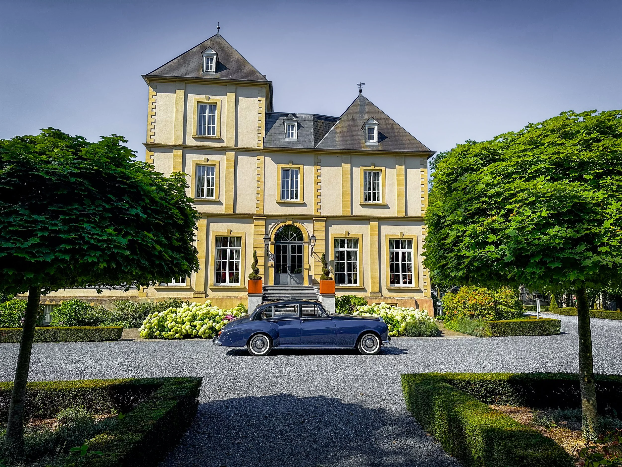 Villa élégante avec un arbre devant, une voiture classique noire garée devant l'entrée, jardins bien entretenus et ciel clair.
