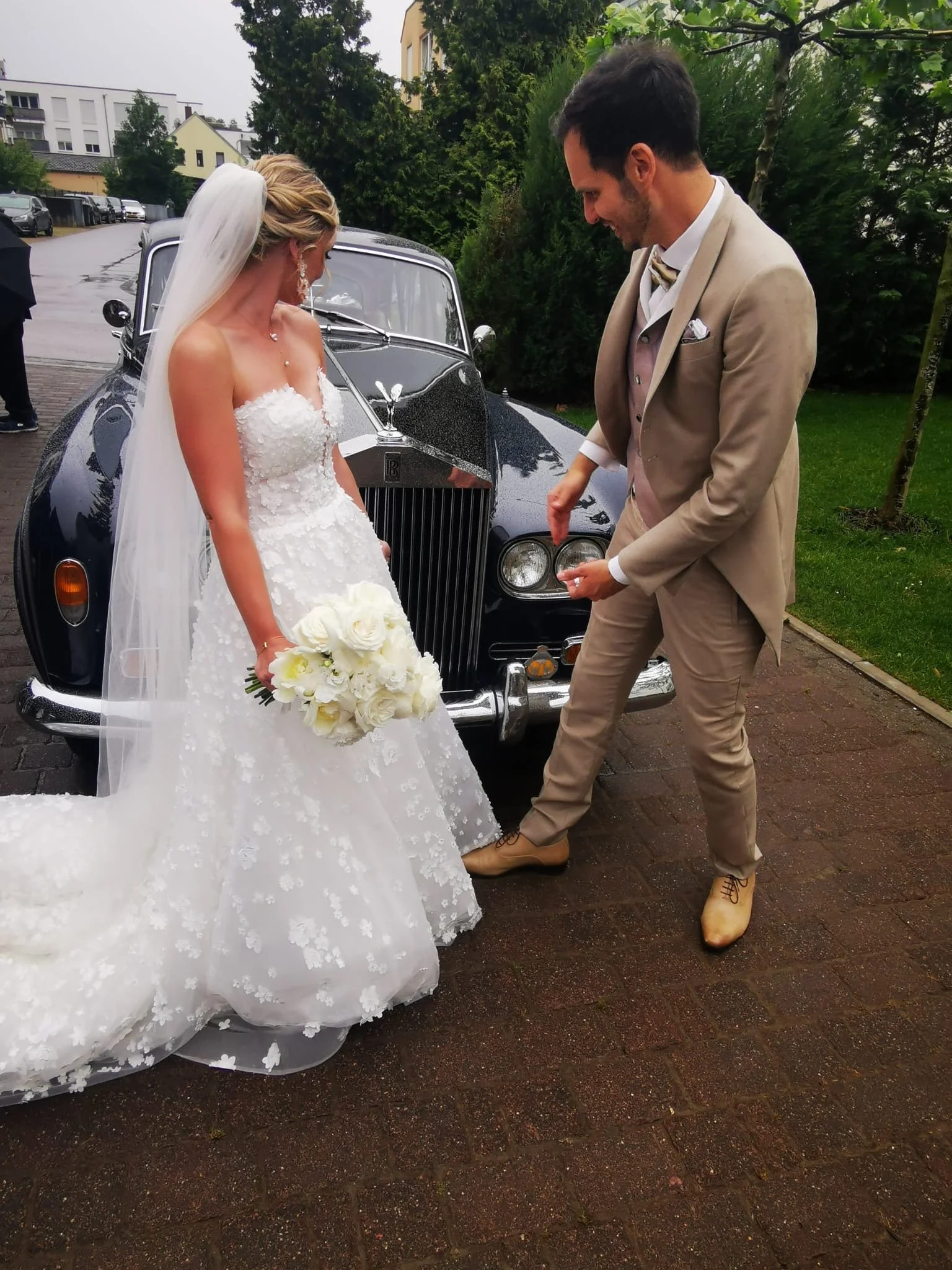 Un couple de mariés en tenue de mariage, la mariée en robe blanche avec bouquet de roses blanches, le marié en costume beige, debout devant une voiture classique noire, probablement lors d'une cérémonie de mariage.