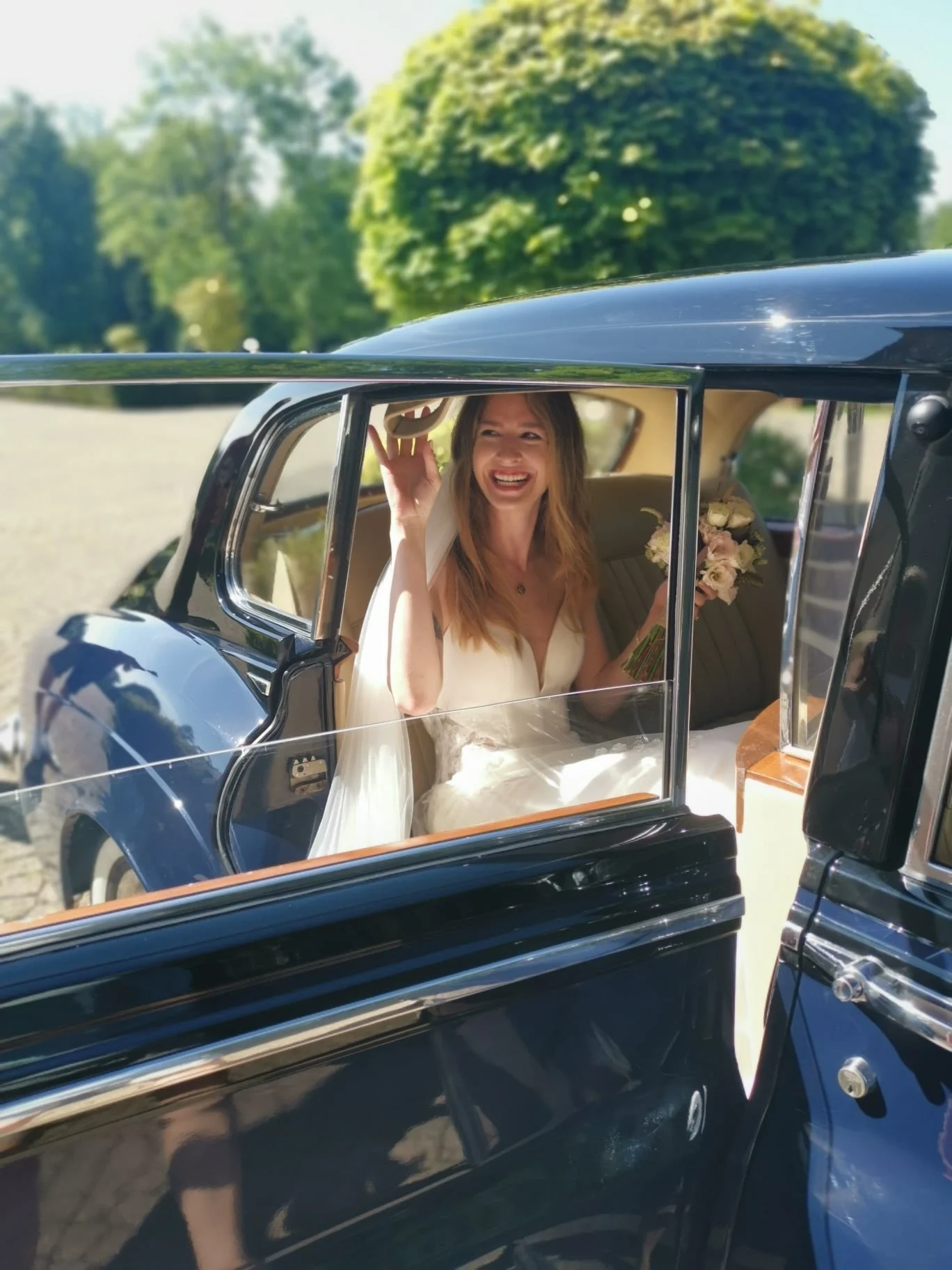 Une femme portant une robe blanche de mariée, assise dans une voiture vintage noire, tenant un bouquet de fleurs, souriant et saluant avec un chapeau beige dans la main.
