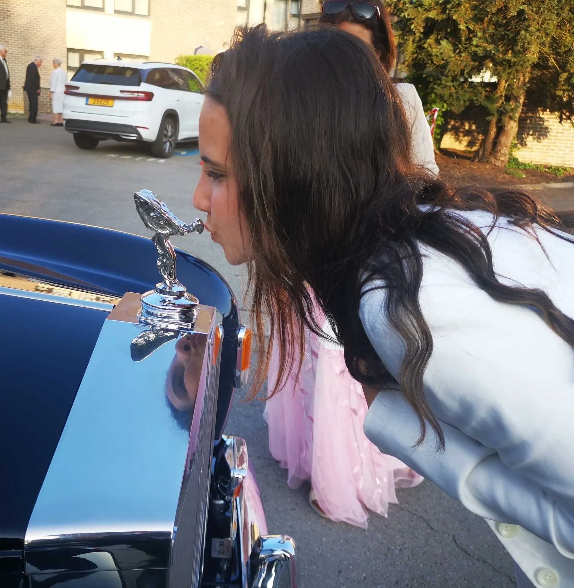 Une femme en train de donner un baiser sur la calandre d'une voiture cabriolet avec un petit trophée argenté, lors d'une sortie en extérieur.