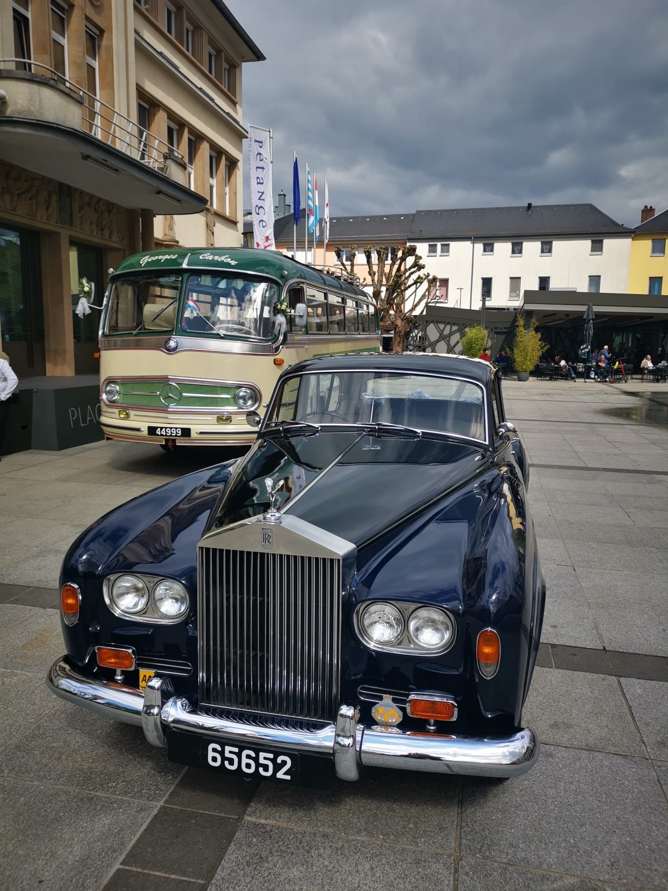 Une voiture classique Rolls-Royce noire et un bus vintage Mercedes-Benz en arrière-plan, exposés sur une place en plein air