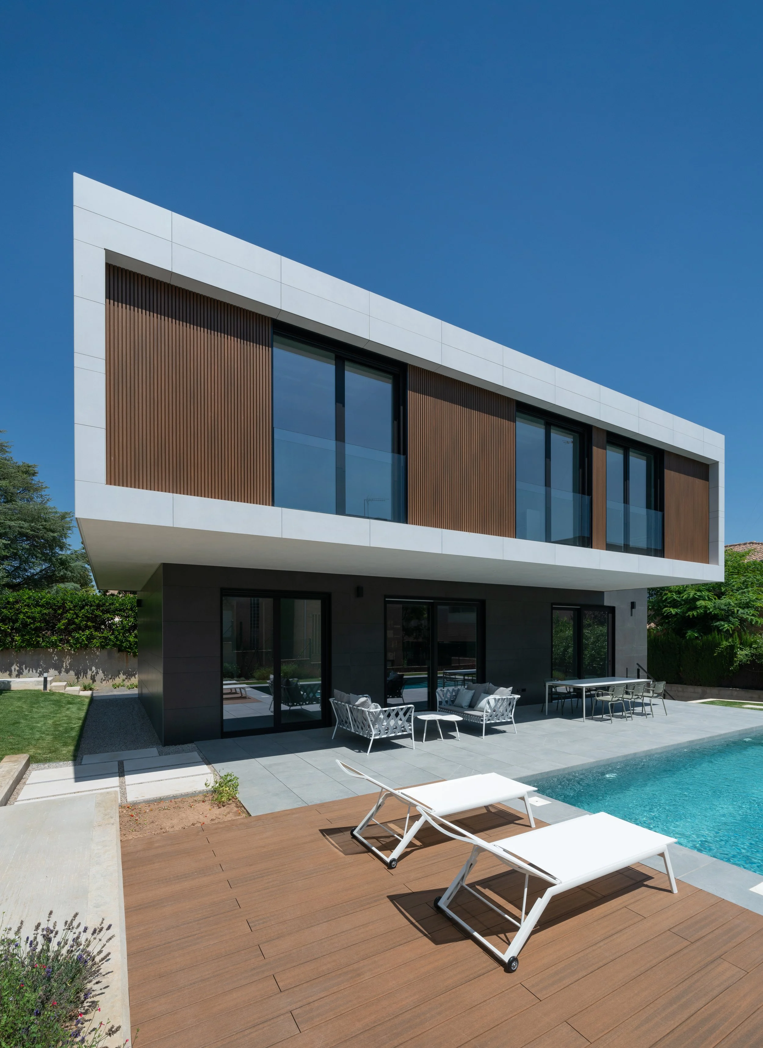 Casa moderna con balcón de vidrio, piscina y muebles exteriores en un día soleado.