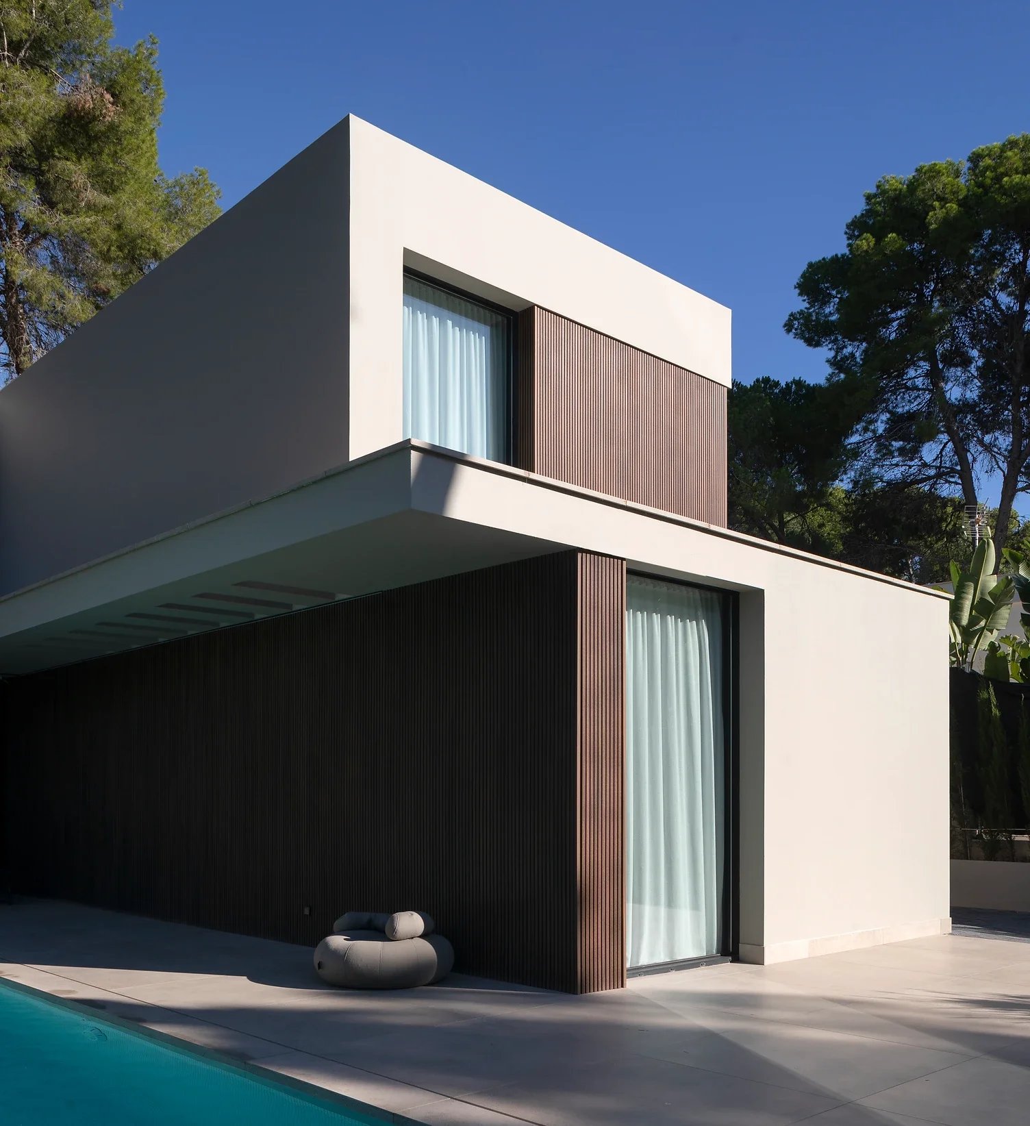 Casa moderna de dos pisos con fachada de paredes blancas y revestimiento de madera compuesta schottendeck, cerca de una piscina y rodeada de árboles verdes tallados, en un día soleado con cielo despejado.