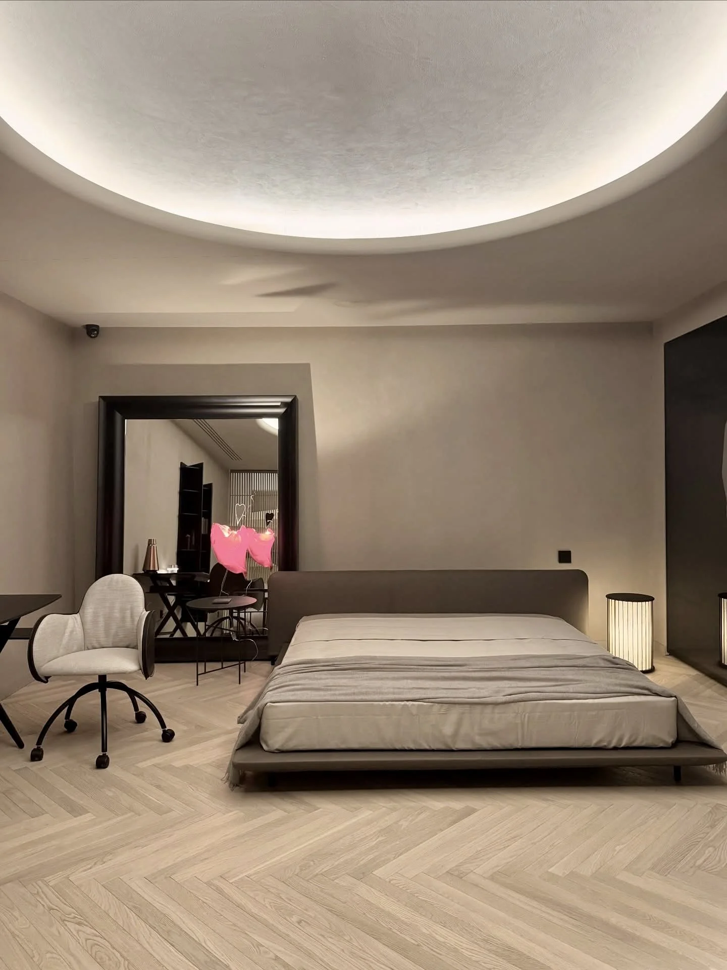 Habitación de lujo con suelo de madera espigua schottenwood con cama baja y colchón, sillón de oficina, espejo grande, lámpara de mesa y decoración minimalista en tonos neutros.