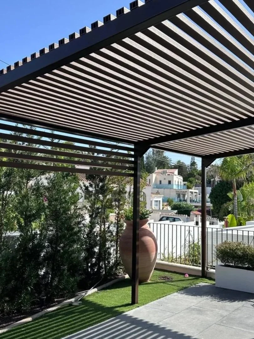 Terraza con pérgola de listones de madera, plantas y macetas decorativas, vista a casas residenciales y árboles en el fondo, cielo azul despejado.