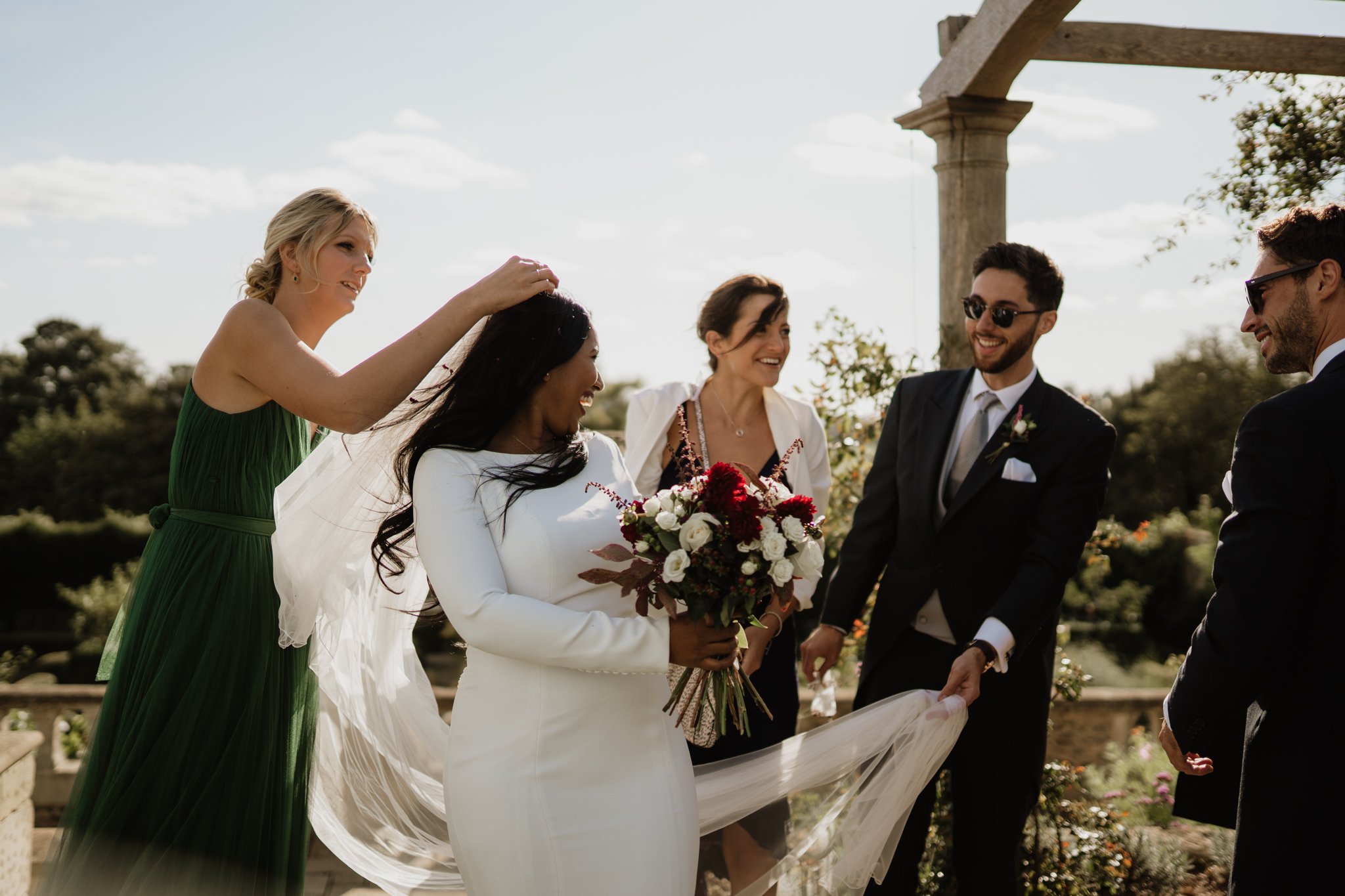 candid photo of bride laughing with groomsmen as bridesmaid reattaches the veil