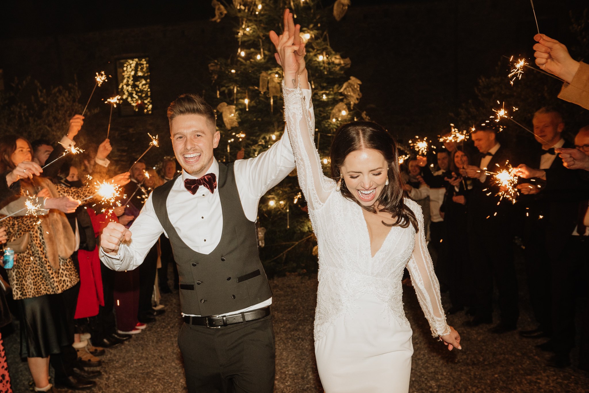 bride and groom cheer as they run through a sparkler tunnel with christmas tree in the background