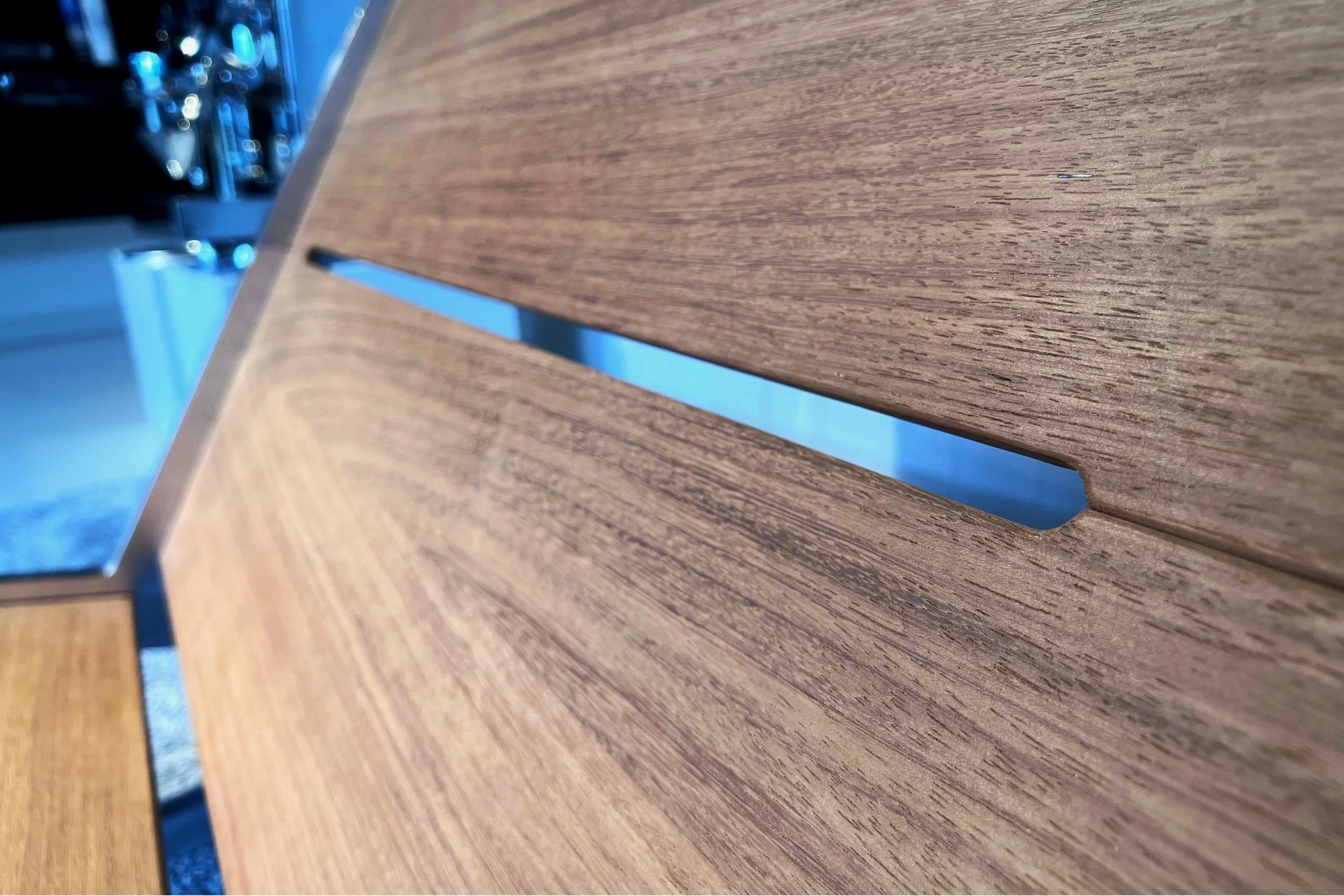 Close-up of a wooden table with a slit or gap in the center, part of a metallic object visible on the left side, and a blue background visible through the gap.