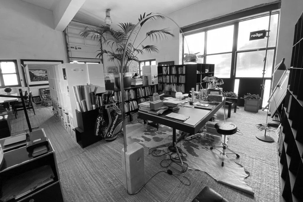 A cluttered home office with a large window letting in natural light, filled with books, papers, plants, and office furniture including a rolling chair, a standing lamp, and various office supplies.