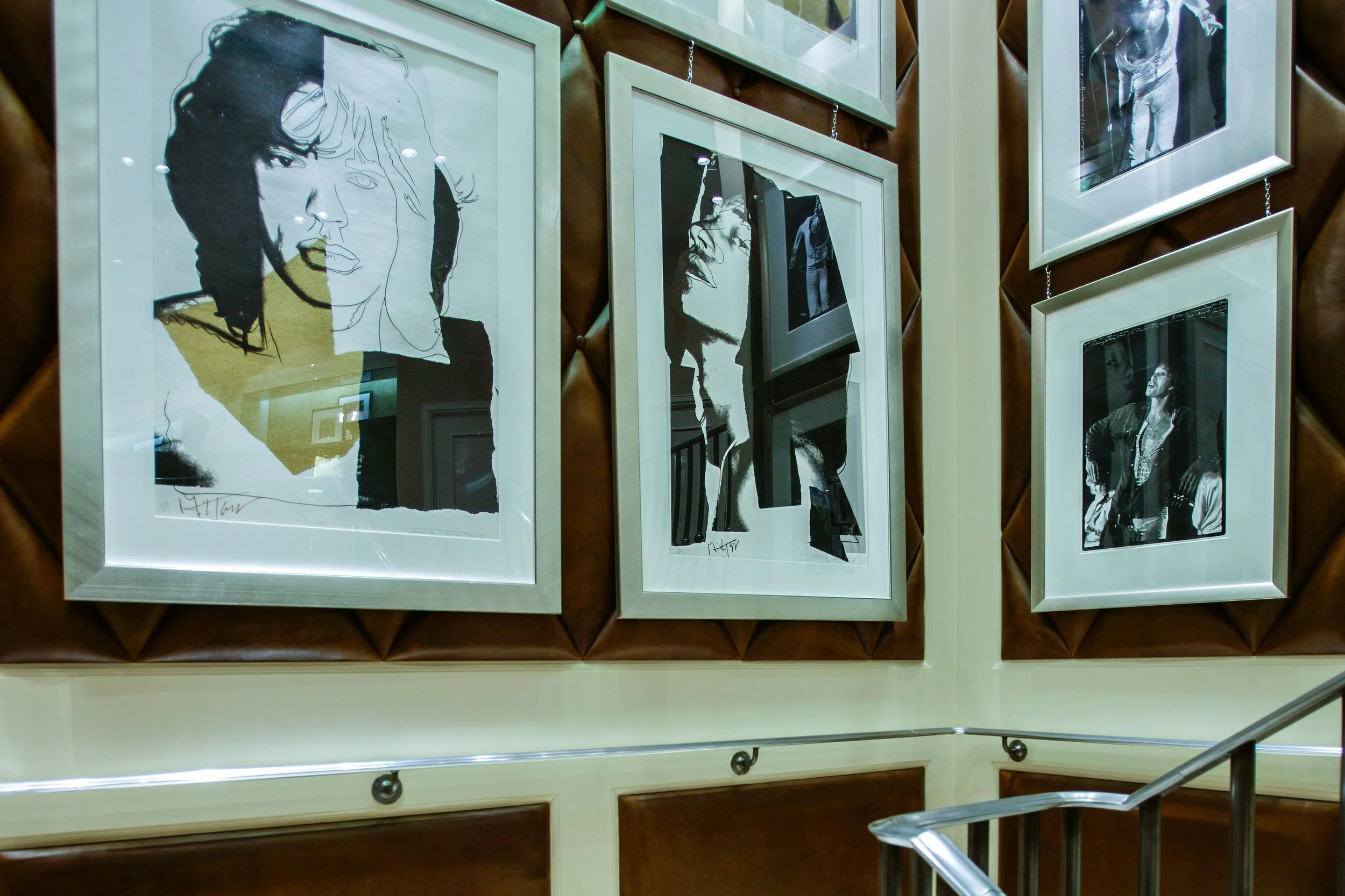 A wall display of framed black-and-white art and photographs, including a line drawing of a woman's face, a profile shot of a woman, and a woman with glasses, in an interior setting with a quilted wall and staircase.
