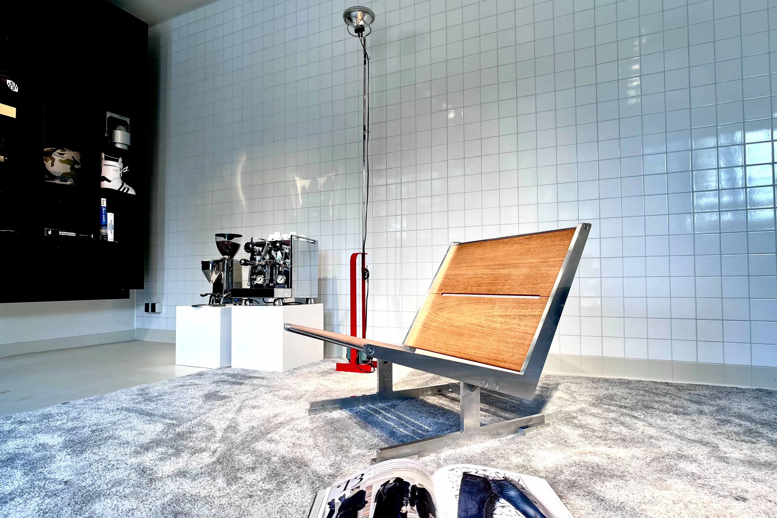 A modern indoor space with white tiled walls, a wooden seating bench, a coffee machine on a white table, and a black shelving unit with various items. The floor is carpeted, and there is a magazine or book open in the foreground.