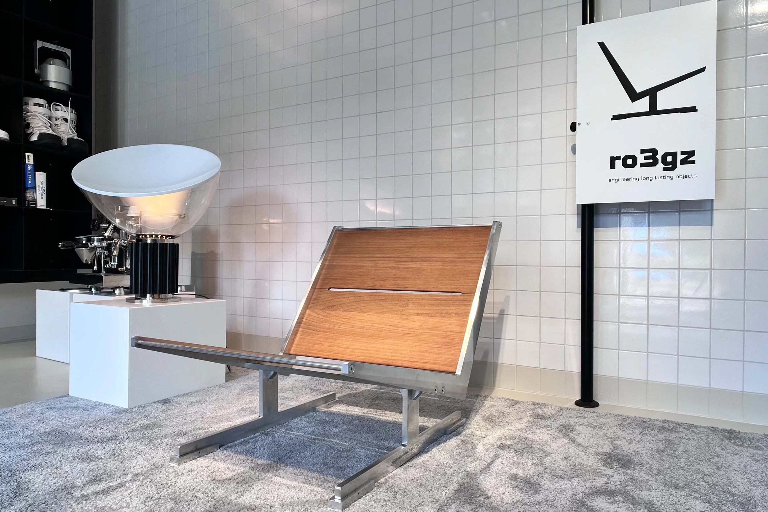 Exhibit in a museum showcasing modern art, including a white curved sculpture on a white pedestal, a wooden bench, a sign with a black abstract design and the text 'ro3gz engineering long lasting objects', and marble tiled walls.