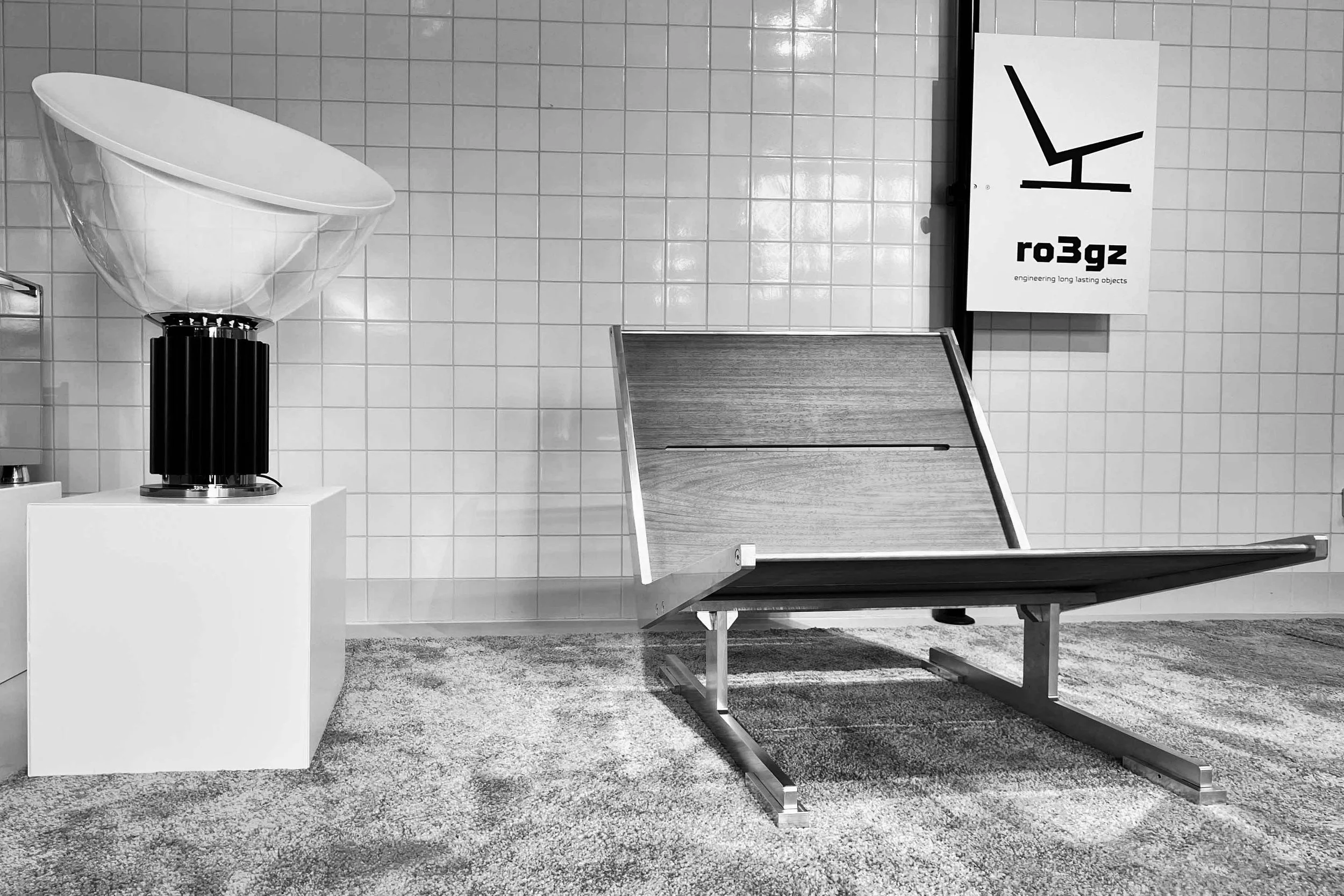 A modern art display featuring a white statue with a large, rounded bowl-shaped top and a black cylindrical base, placed on a white pedestal. Beside it is an empty wooden inclined desk or table, and in the background, a sign with a logo and the text "ro3gz engineering long lasting objects" is mounted on a tiled wall.