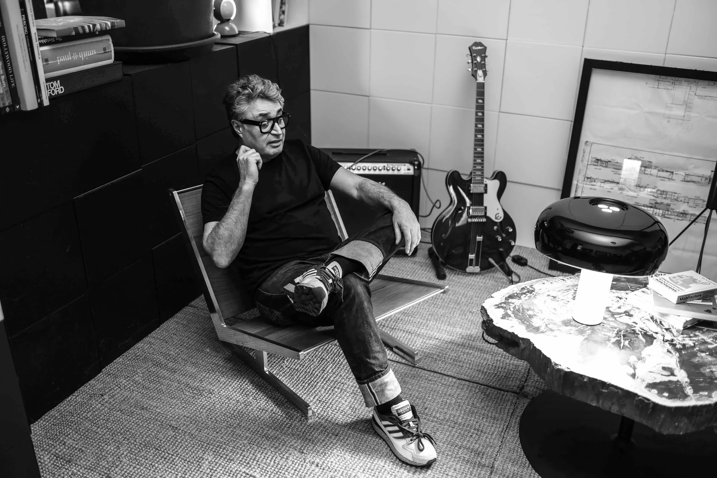 Black and white photo of a man sitting on a chair in a room with musical instruments, books, and modern decor. He is wearing glasses, a black shirt, jeans, and sneakers, and is holding his chin in thought.