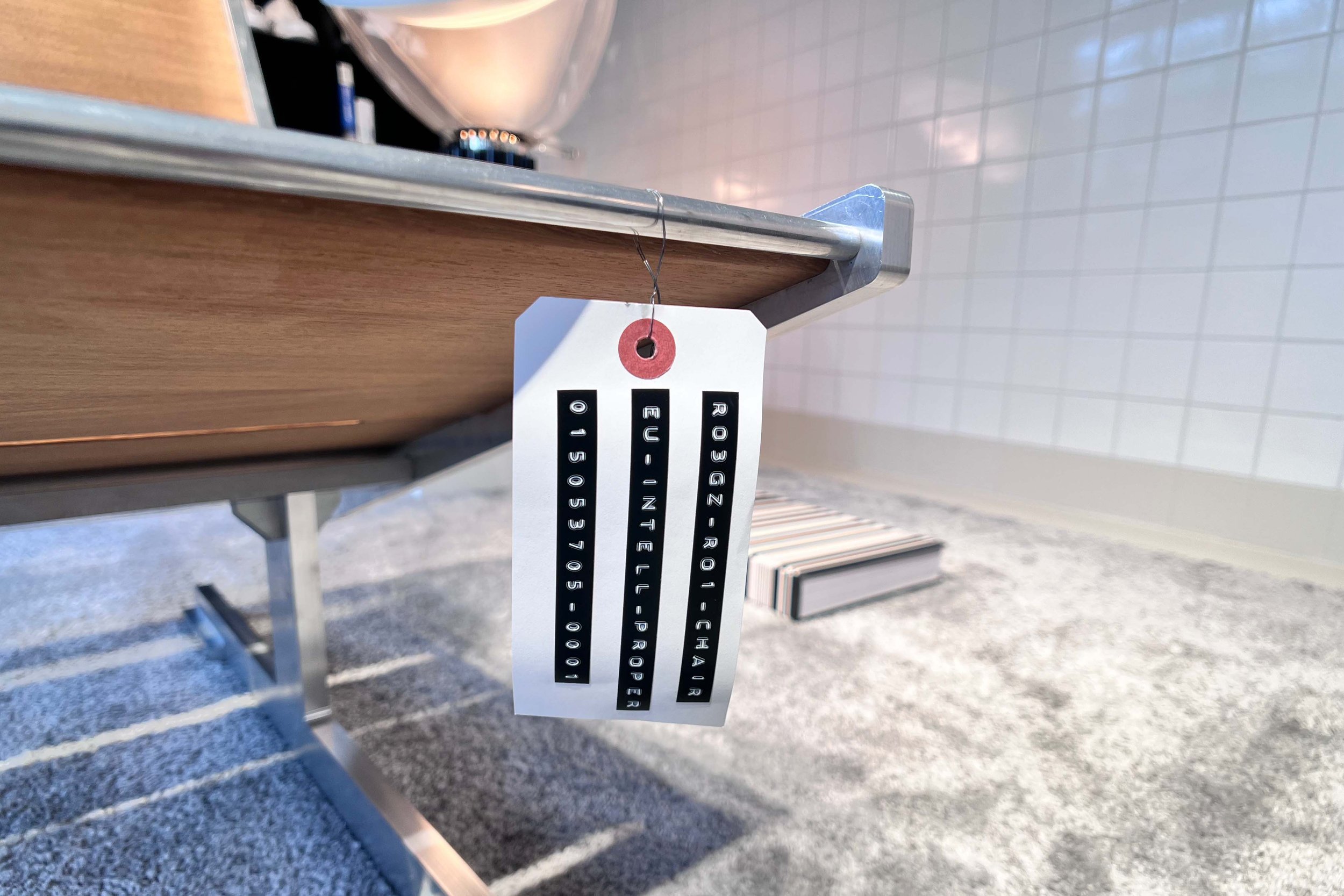 A close-up of a white and black label with a red hole reinforcement, hanging from a metal edge of a wooden surface, with a blurred background of a tiled wall and a concrete floor.