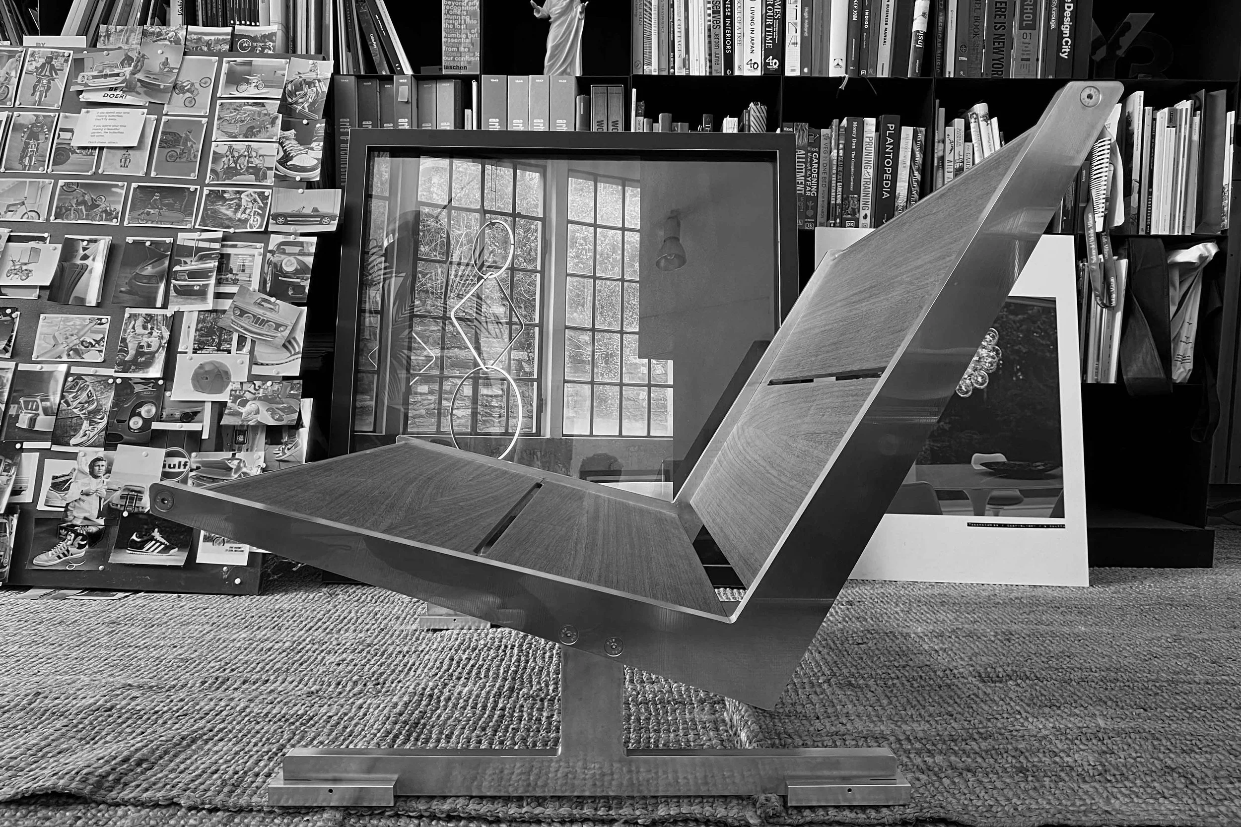A modern wooden coffee table with angled legs in a room with a large window and bookshelves. The bookshelves contain books, magazines, and decorative items. The wall behind the shelves is decorated with photos and postcards. A mirror is leaning against the wall, reflecting the window and trees outside.