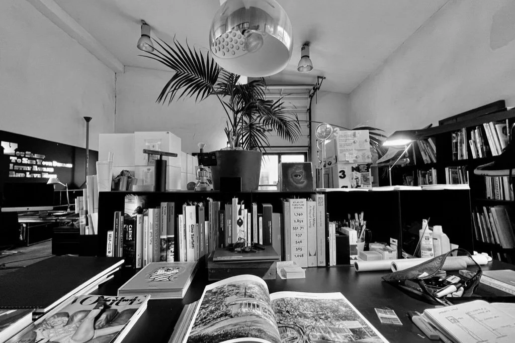 A cluttered office or studio space with bookshelves filled with books, a large potted plant, a desk with open magazines, and various office supplies. There is a window in the background and a large ceiling light.