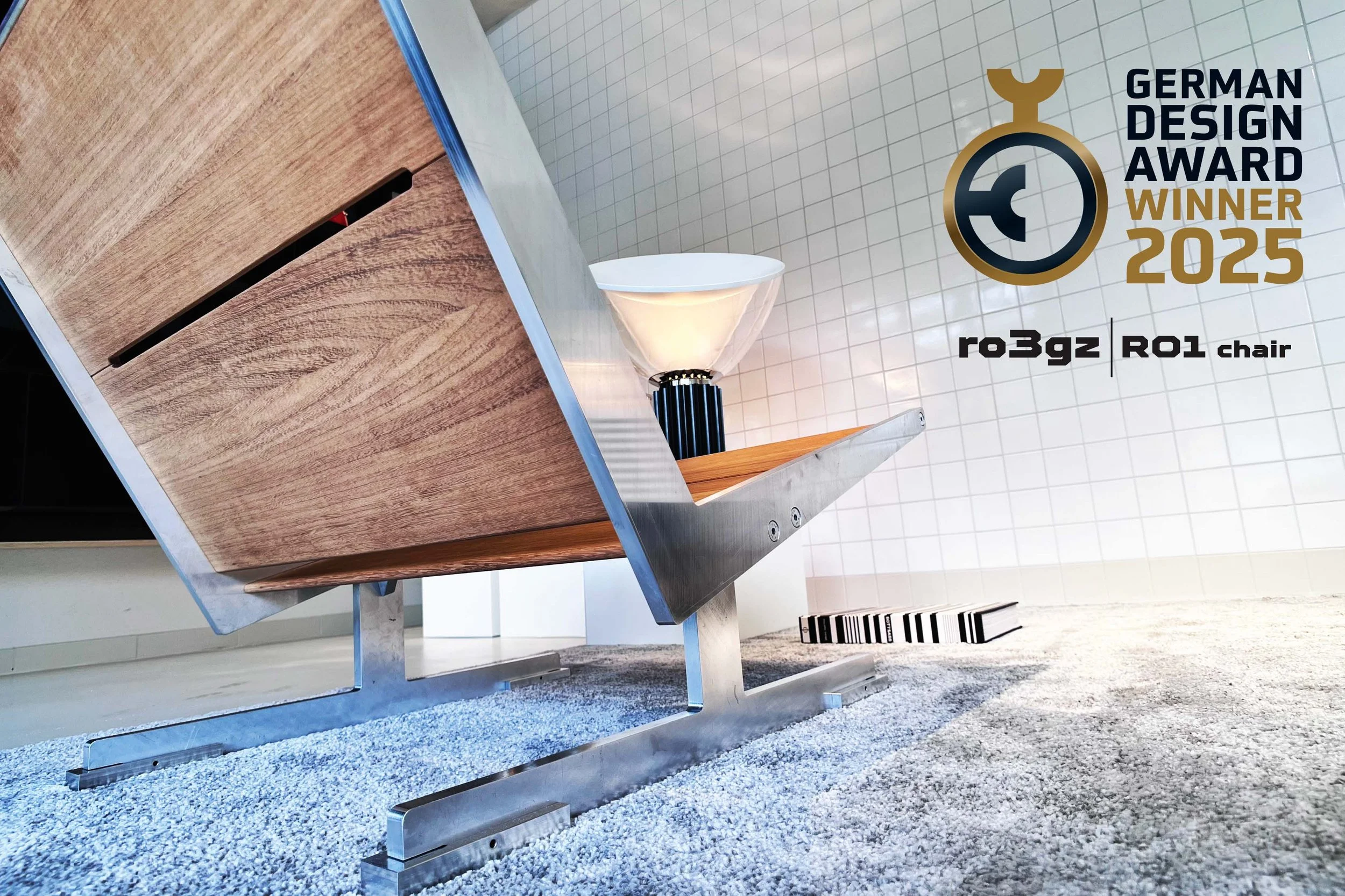 Modern wooden and metal chair with a white bowl on top, set on a gray textured surface, with a tiled wall displaying a German design award for 2025.