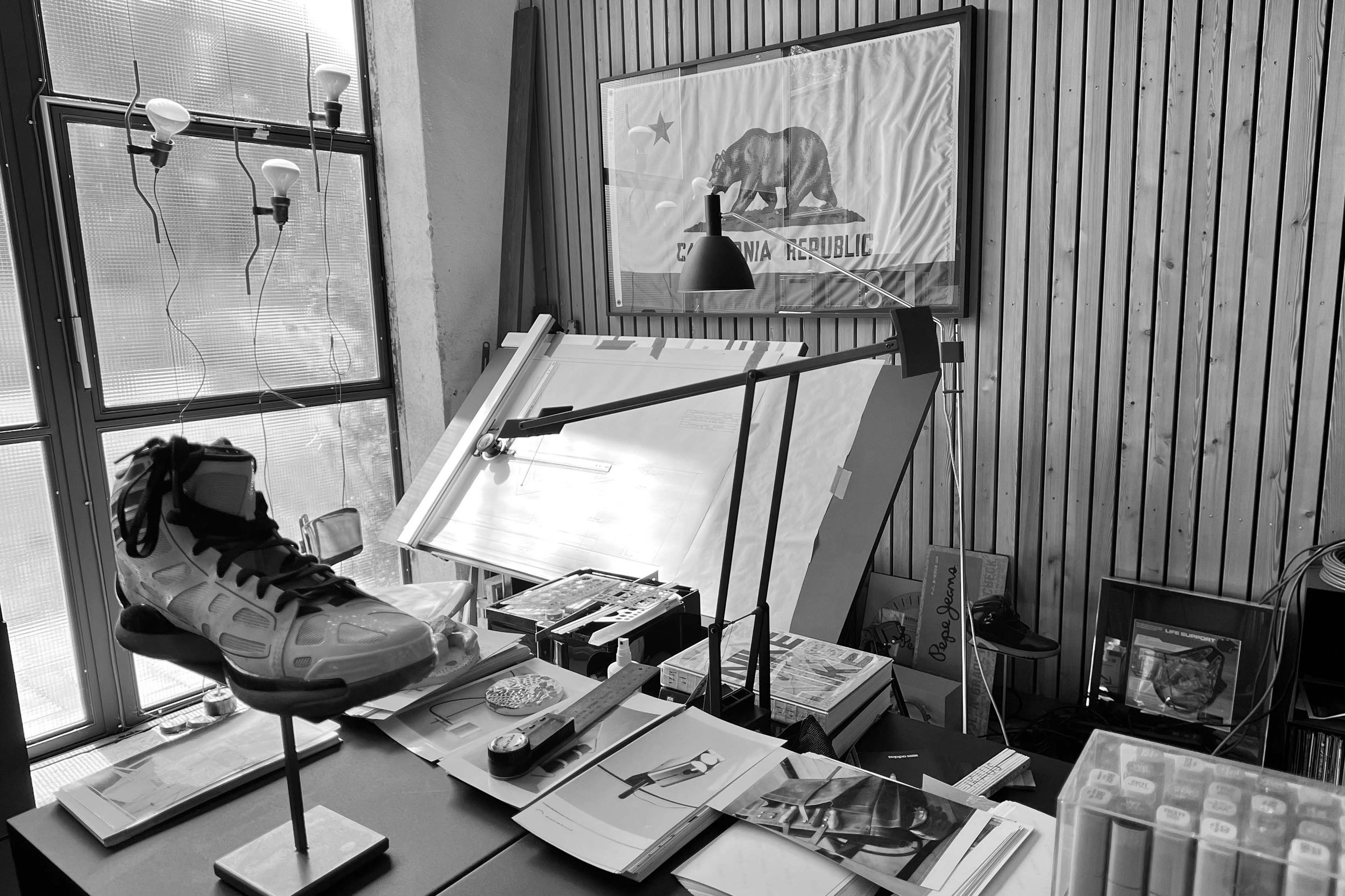 A workspace with various office supplies, a model of a foot and ankle brace, and a drafting table with papers. There is a window with hanging light bulbs and a California Republic flag on the wall.