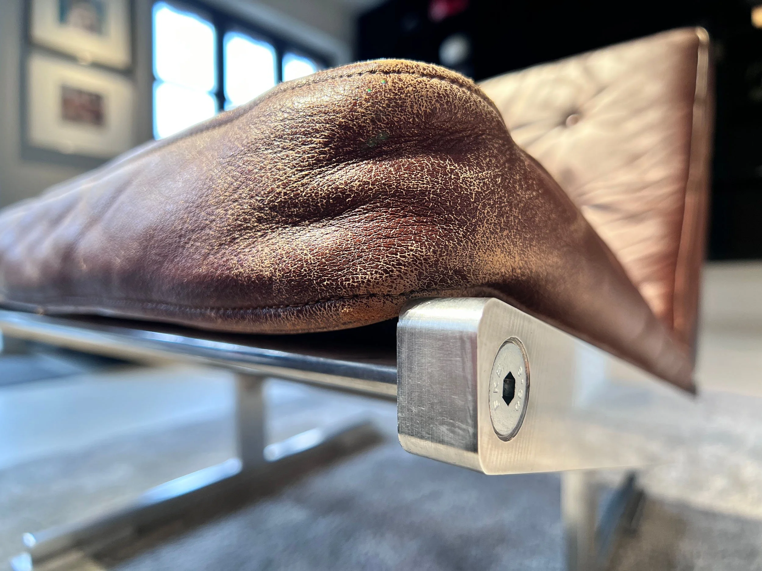 Close-up of a brown leather shoe with detailed stitching, resting on a metallic surface, with a blurred background featuring windows and picture frames.