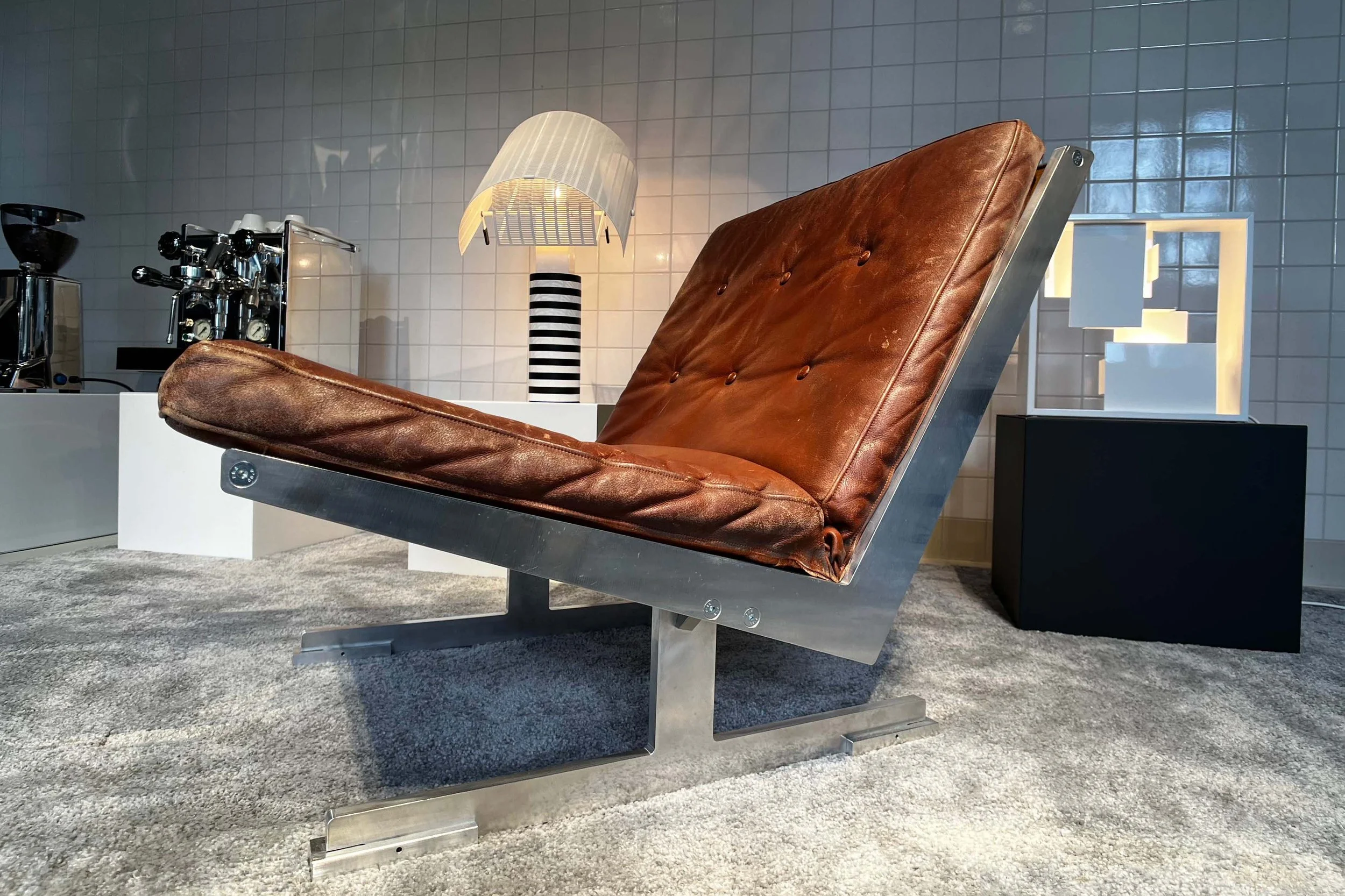 Mid-century modern lounge chair with leather upholstery and metal frame, positioned on a carpeted floor against a tiled wall. In the background, a lamp with a striped base and a coffee machine are visible.