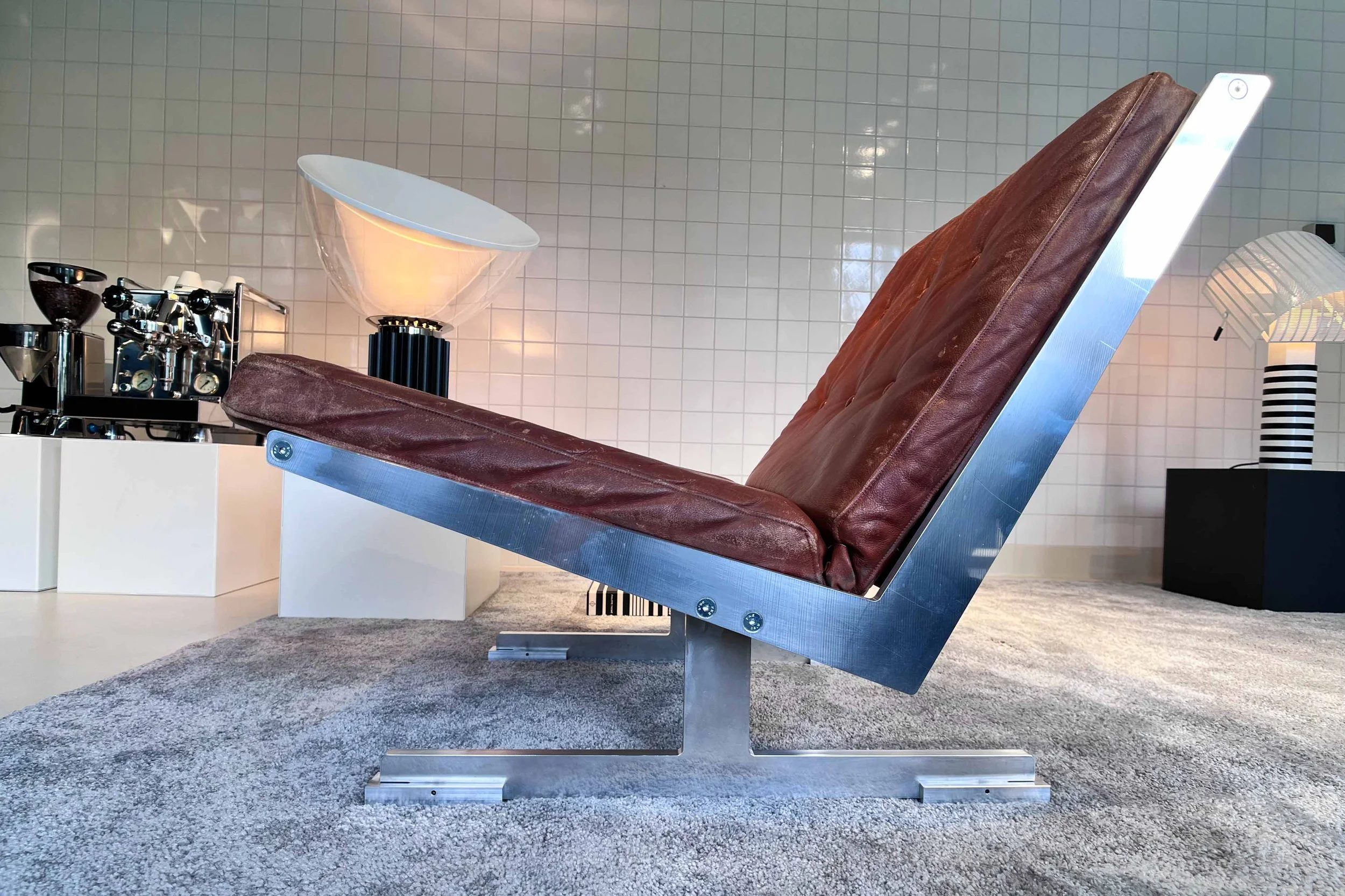 A vintage-style reclining lounge chair with a brown leather cushion and metallic frame, situated on a gray carpet in an indoor space with white tiled walls, alongside lamps and modern decor.
