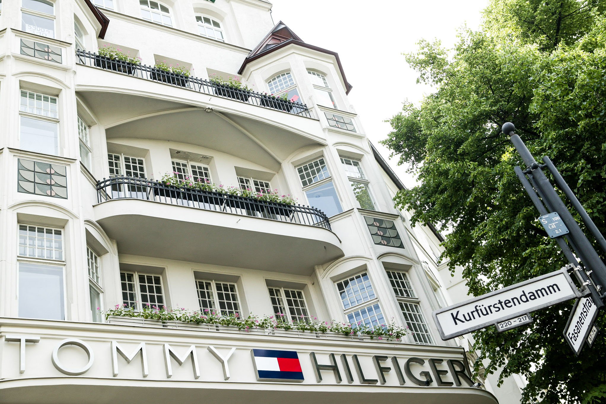 A multi-story white building with ornate architectural details, balconies with black railings and pink flowers, and large windows. A street sign in the foreground indicates the intersection of Kurfürsten(damm) and other streets. A green tree partially obscures the right side of the building.