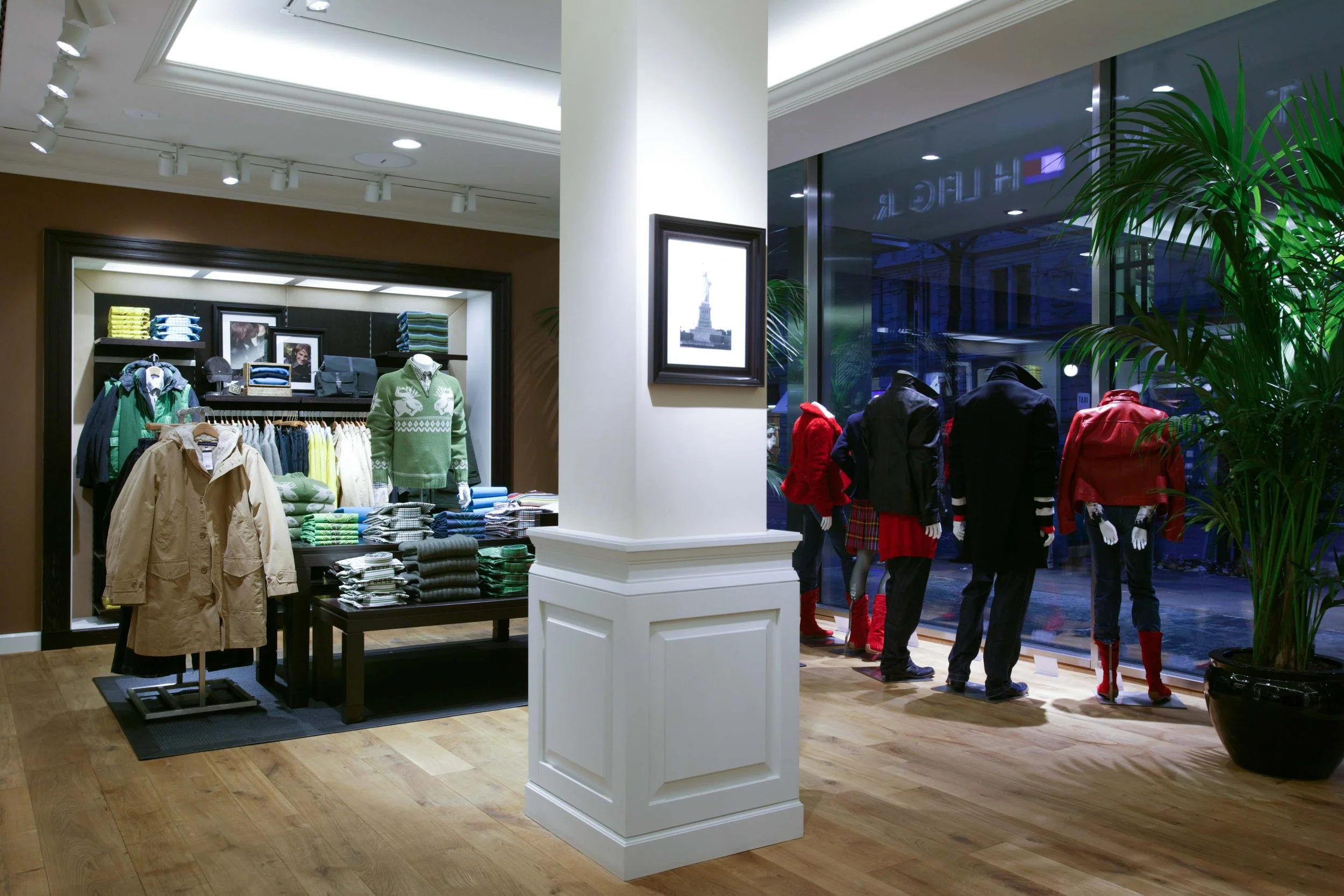 A clothing store window display featuring mannequins dressed in colorful winter apparel, with a large potted plant and framed artwork inside.