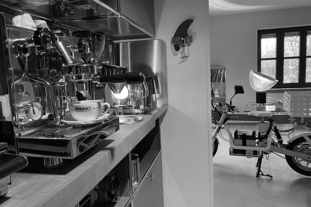 A kitchen with coffee-making equipment, a cup on a saucer labeled 'ROCKET', and a partially visible wall-mounted clock on the left. In the background, a room with a bicycle, a lamp, and a window with natural light.
