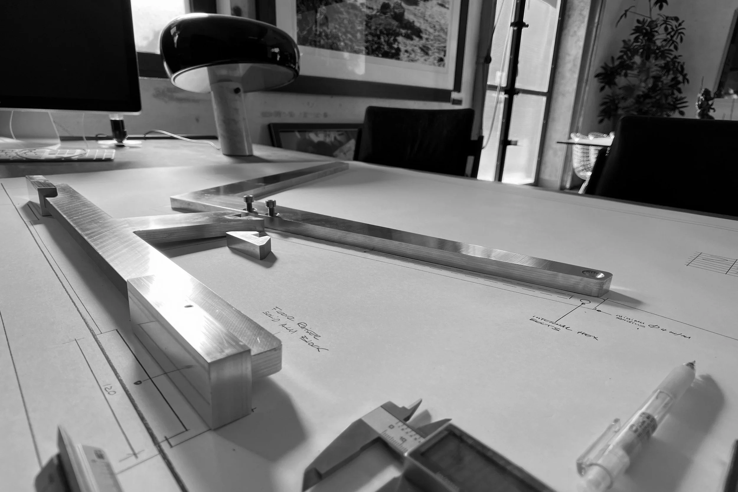A drafting table with architectural plans, a ruler, and metal framing pieces, set in a room with large windows, a computer monitor, a desk lamp, and a potted plant.
