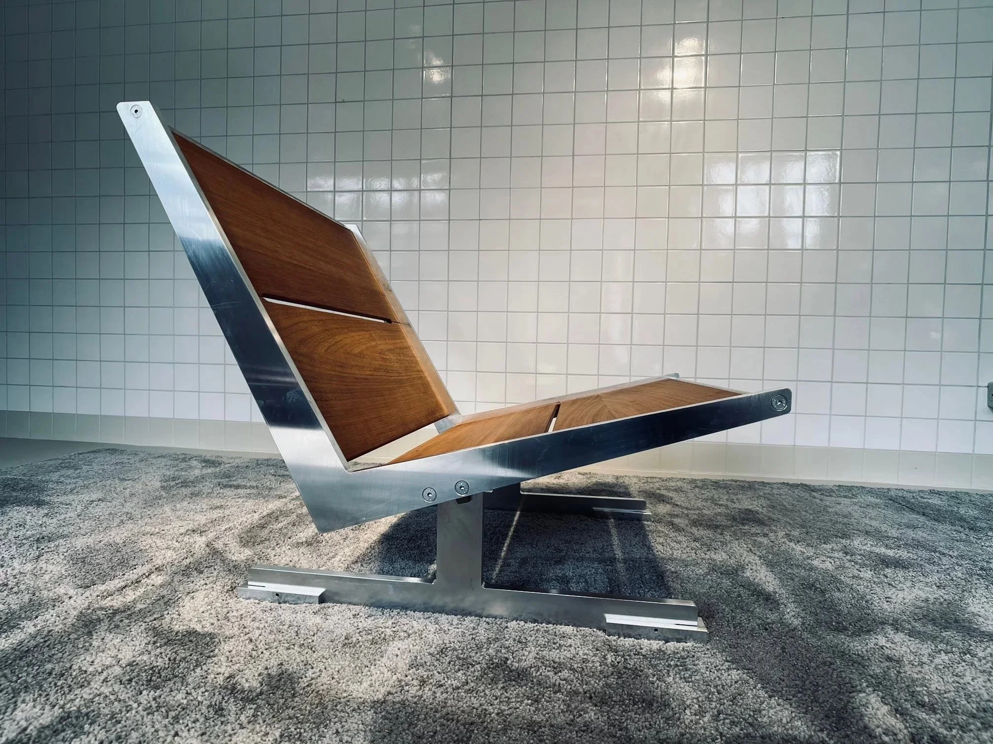 Modern wooden and metal chair with a slanted backrest, situated against a tiled wall and gray carpeted floor.