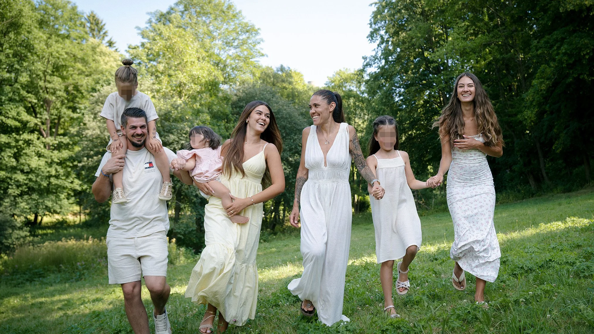 Eine glückliche Gruppe von sieben Menschen, darunter Kinder und Erwachsene, spaziert lächelnd durch einen grünen Park mit Bäumen an einem sonnigen Tag. Entstanden bei einem Familien Shooting in Schmitten im Taunus.