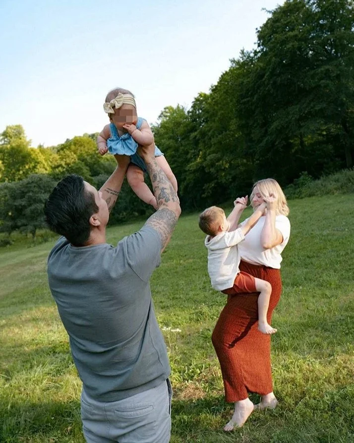 Ein Mann hebt ein Baby in die Luft, während eine Frau und ein Junge auf einer Wiese spielen, im Hintergrund Bäume und ein blauer Himmel. Entstanden bei einem Familien Shooting in Schmitten im Taunus.